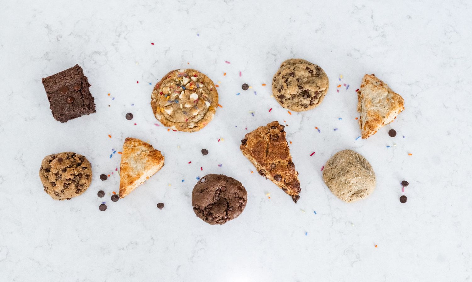 Assorted baked goods including chocolate chip cookies, brownies, and scones arranged on a white marble surface.