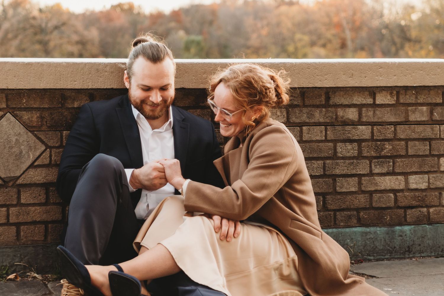 Laughing couple sits against a brick wall outdoors surrounded by colorful autumn foliage.