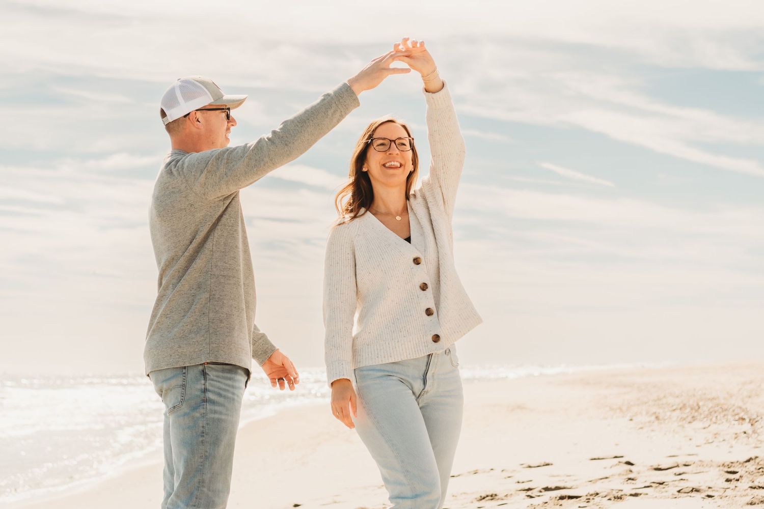 Happy couple dances playfully on a sunny beach with blue skies and ocean waves behind them.
