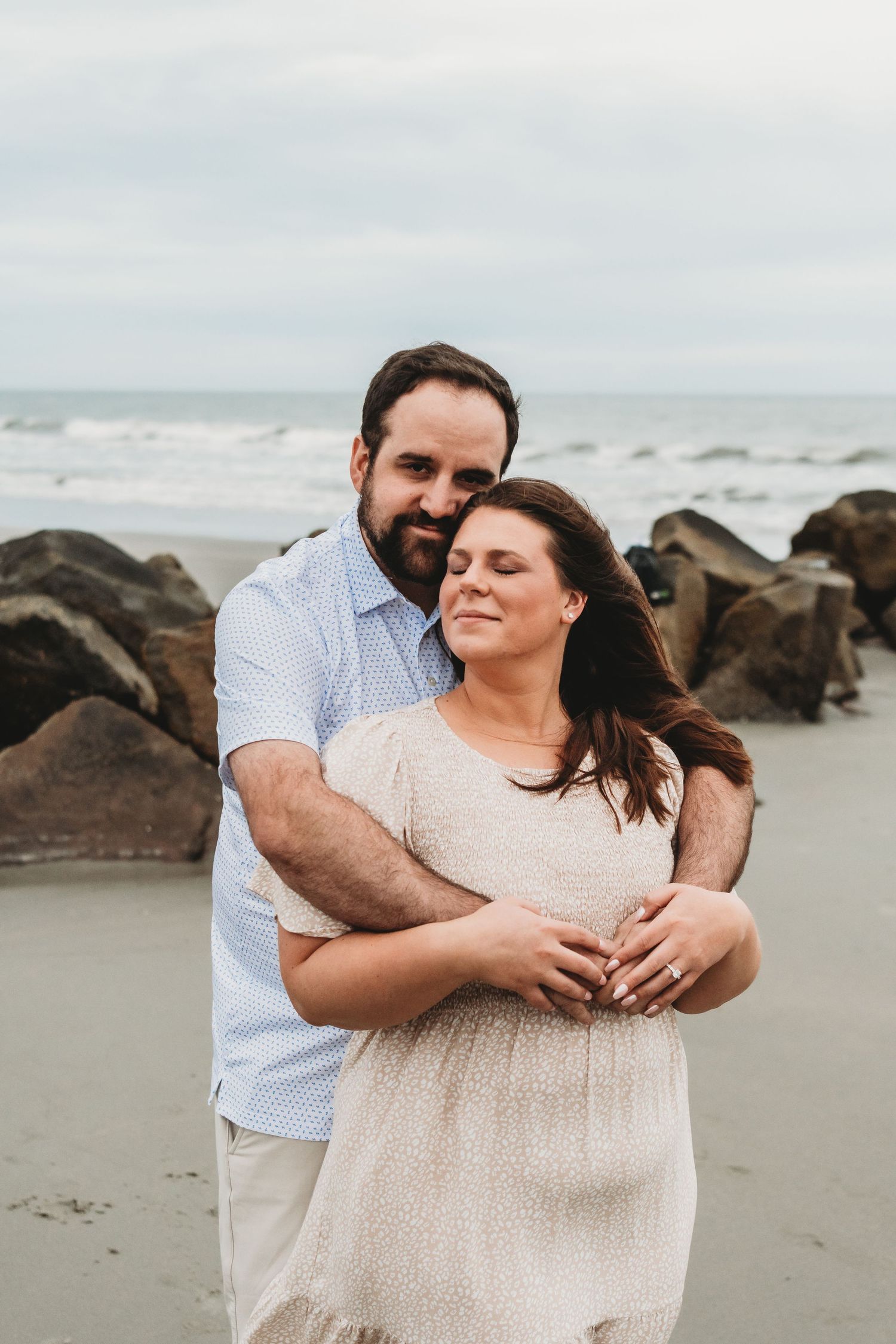A bearded man tenderly embraces a smiling woman from behind on a rocky beach with ocean waves in the background.