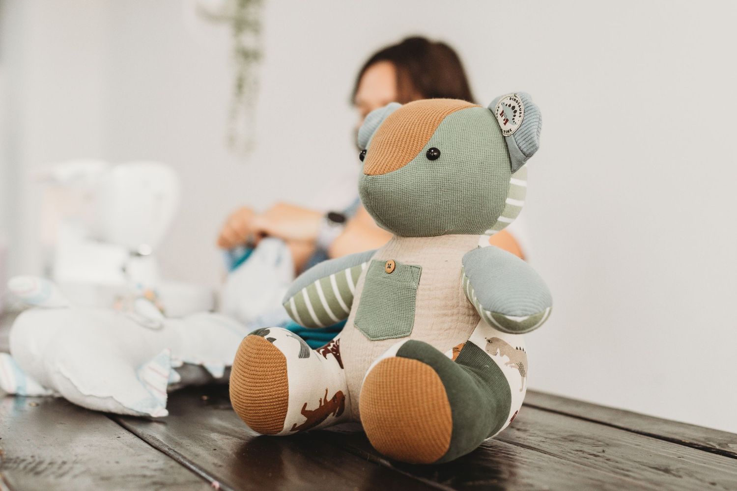 A colorful patchwork teddy bear sits on a wooden floor with a blurred woman and baby items in the background.