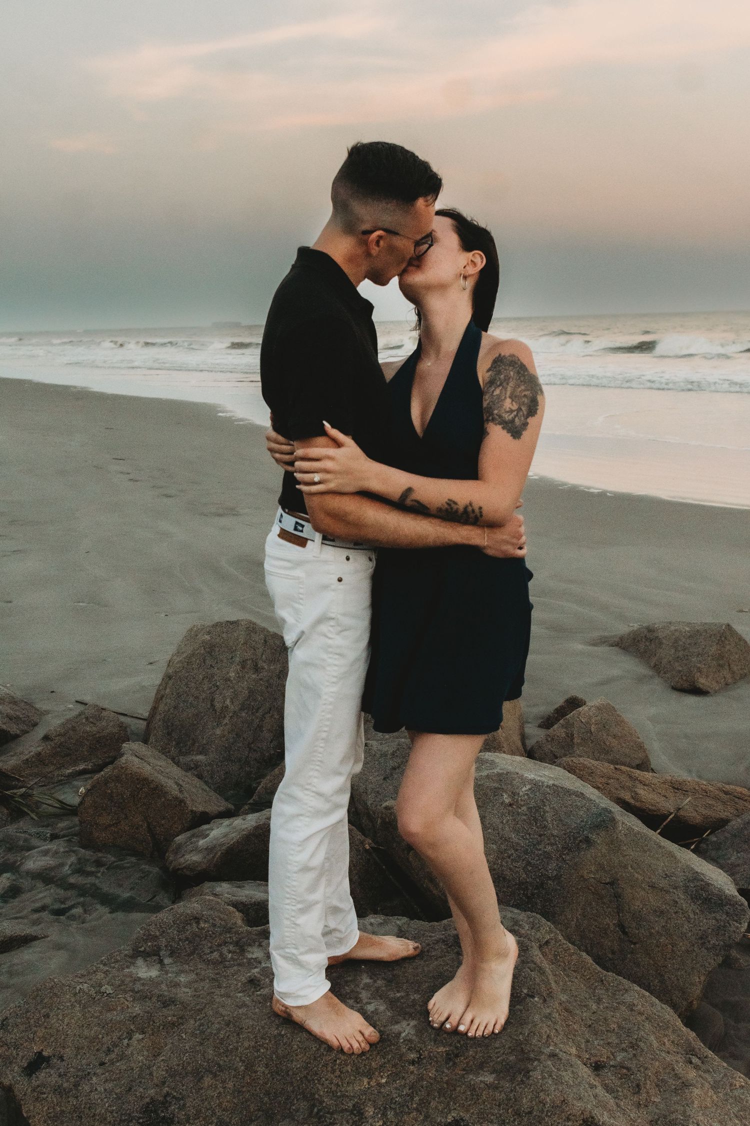A couple sharing a passionate kiss while standing on rocky shoreline at the beach during golden hour.