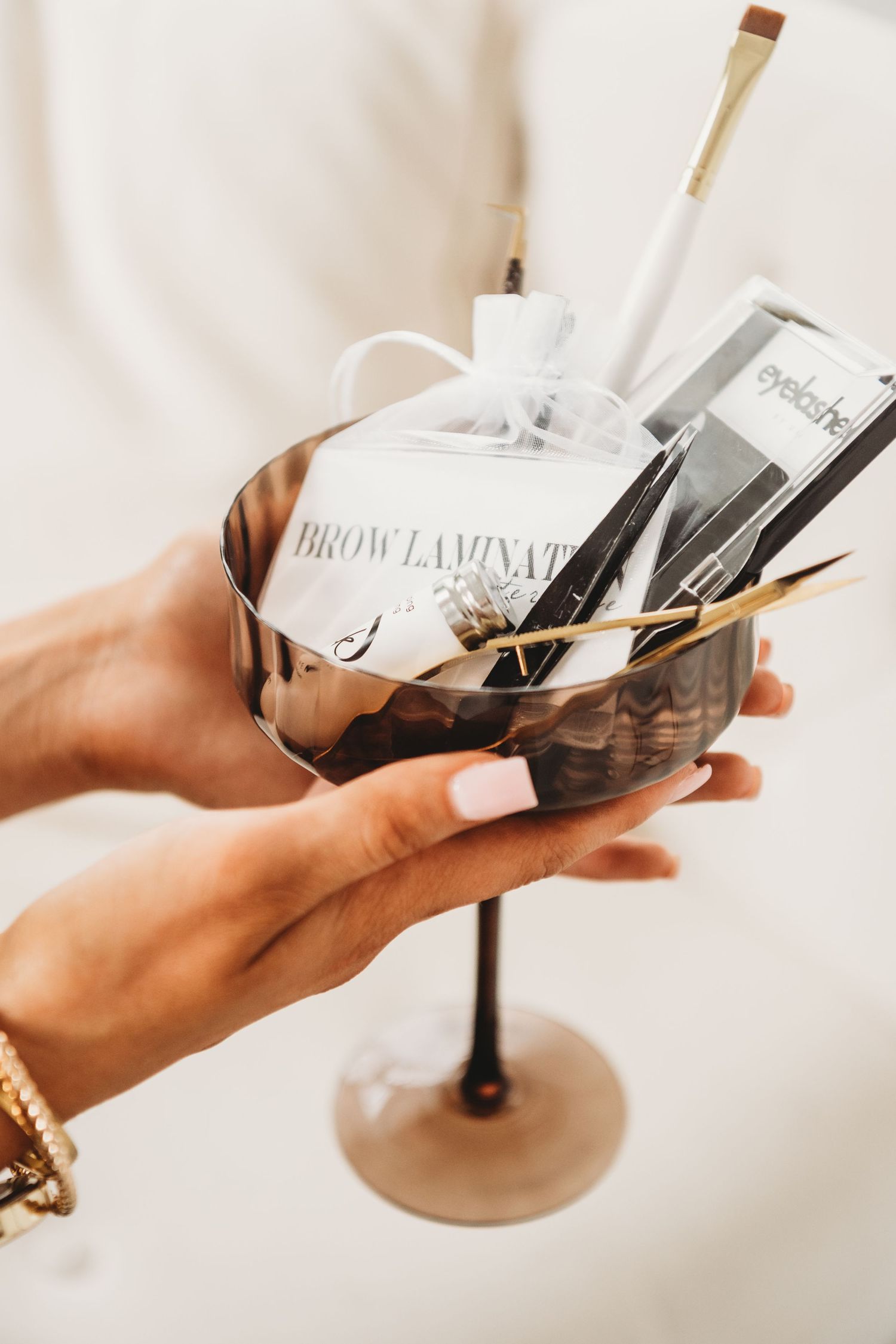 Hand holding decorative martini glass filled with brow lamination beauty tools.