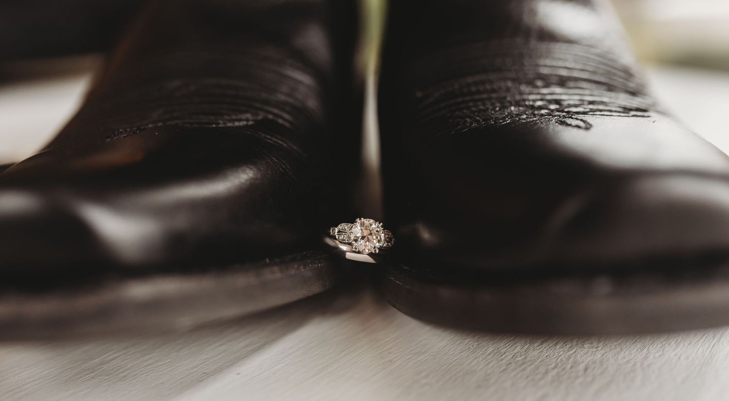 Diamond engagement ring nestled between polished dark leather dress shoes on light floor.