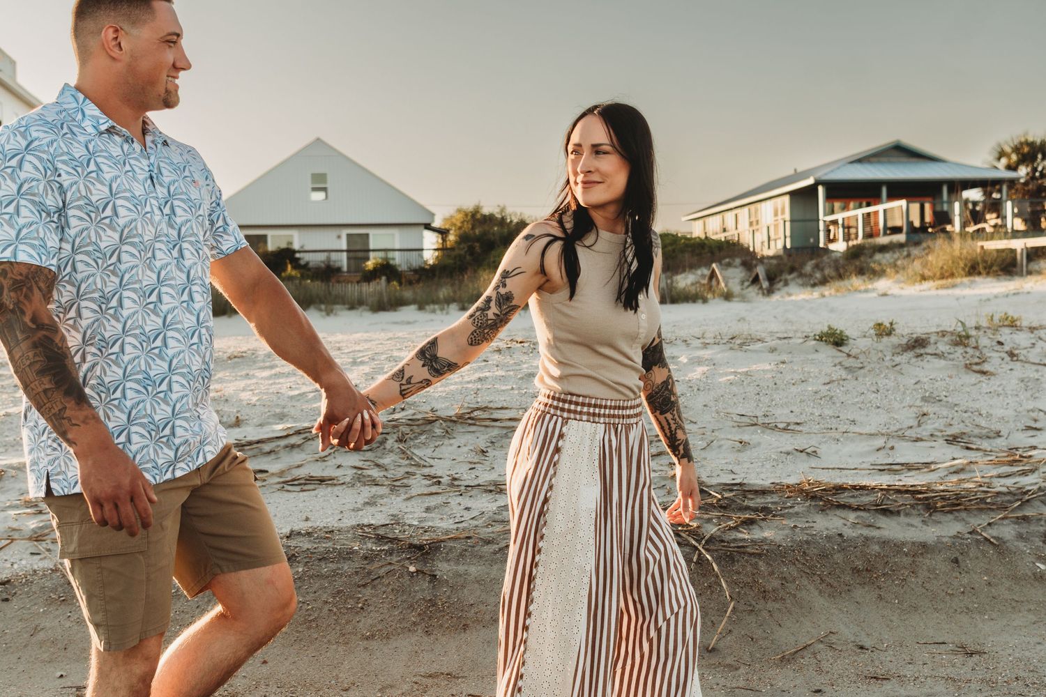 Couple holding hands walking on sandy beach near coastal homes at sunset.