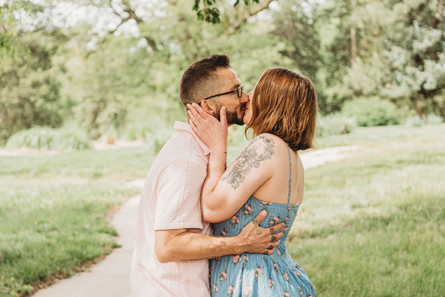 A couple sharing a kiss on a garden path, woman in a blue floral dress with a shoulder tattoo.