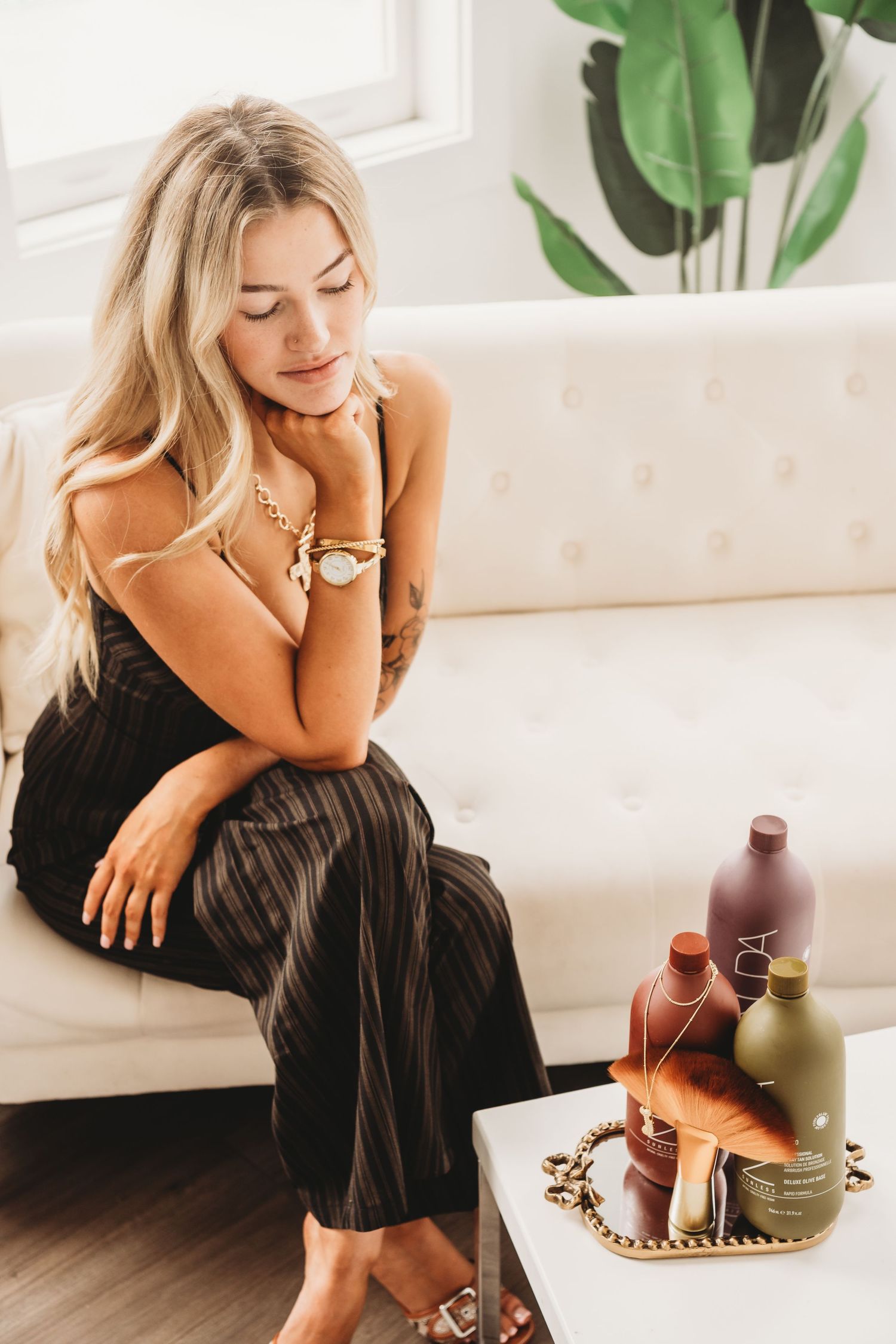 Blonde woman in black striped jumpsuit sitting on white couch near beauty products.