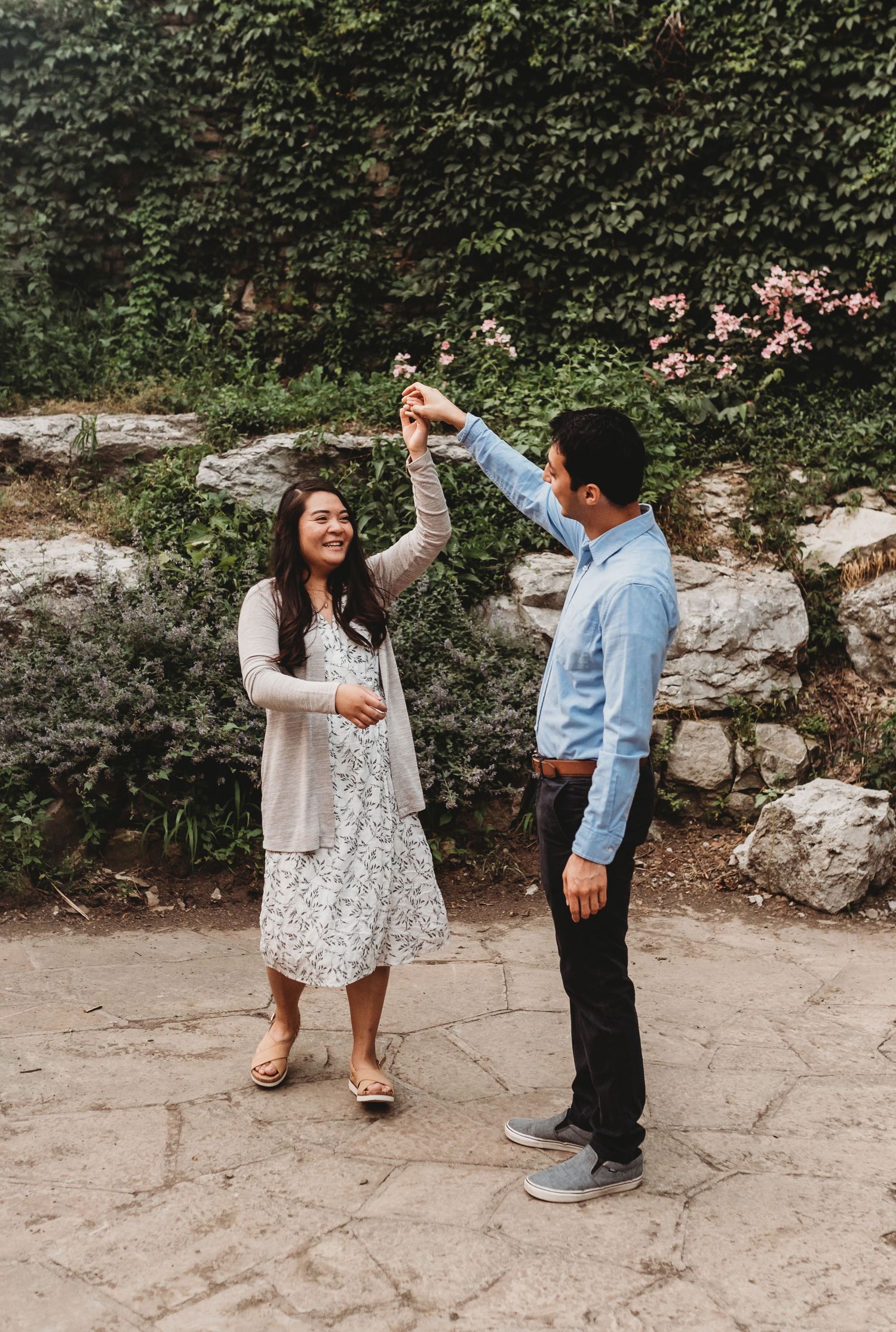 A joyful couple dances outdoors near a stone wall with blooming flowers and green ivy.
