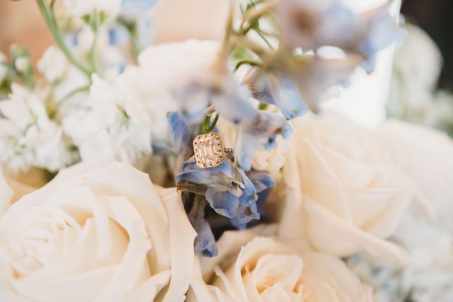 Close-up of a halo diamond engagement ring resting on white roses and blue flowers in a bouquet.