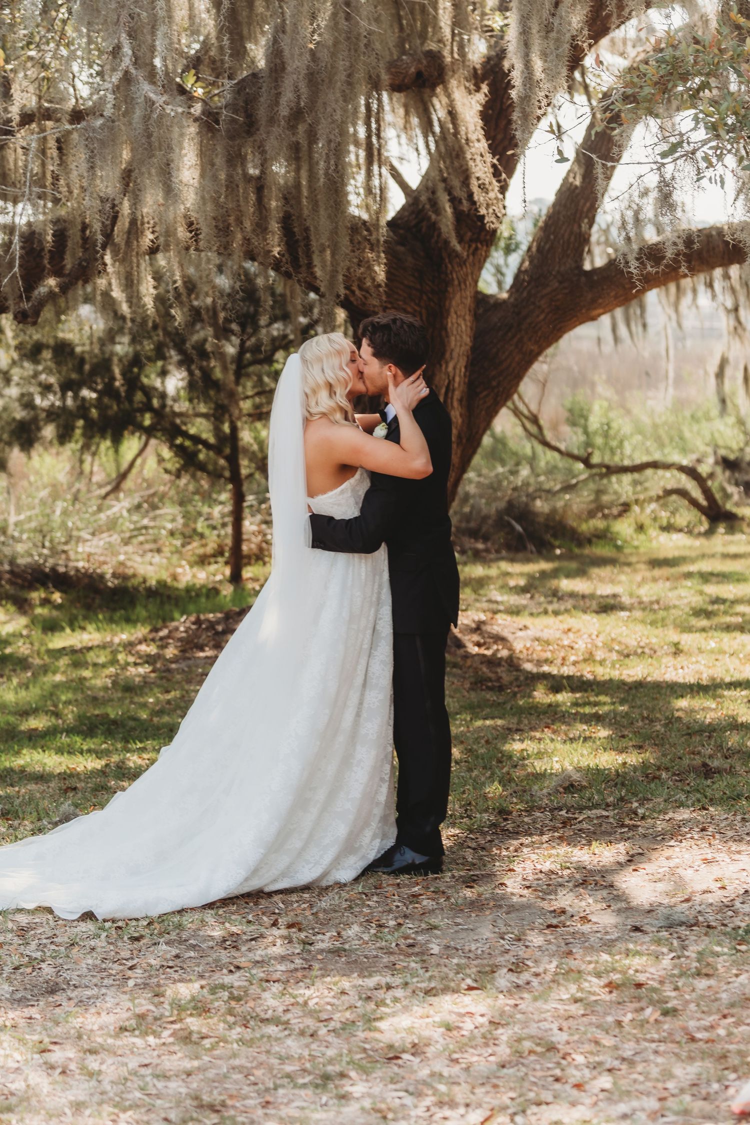 Bride and groom share a romantic kiss under a moss-draped oak tree on their wedding day.