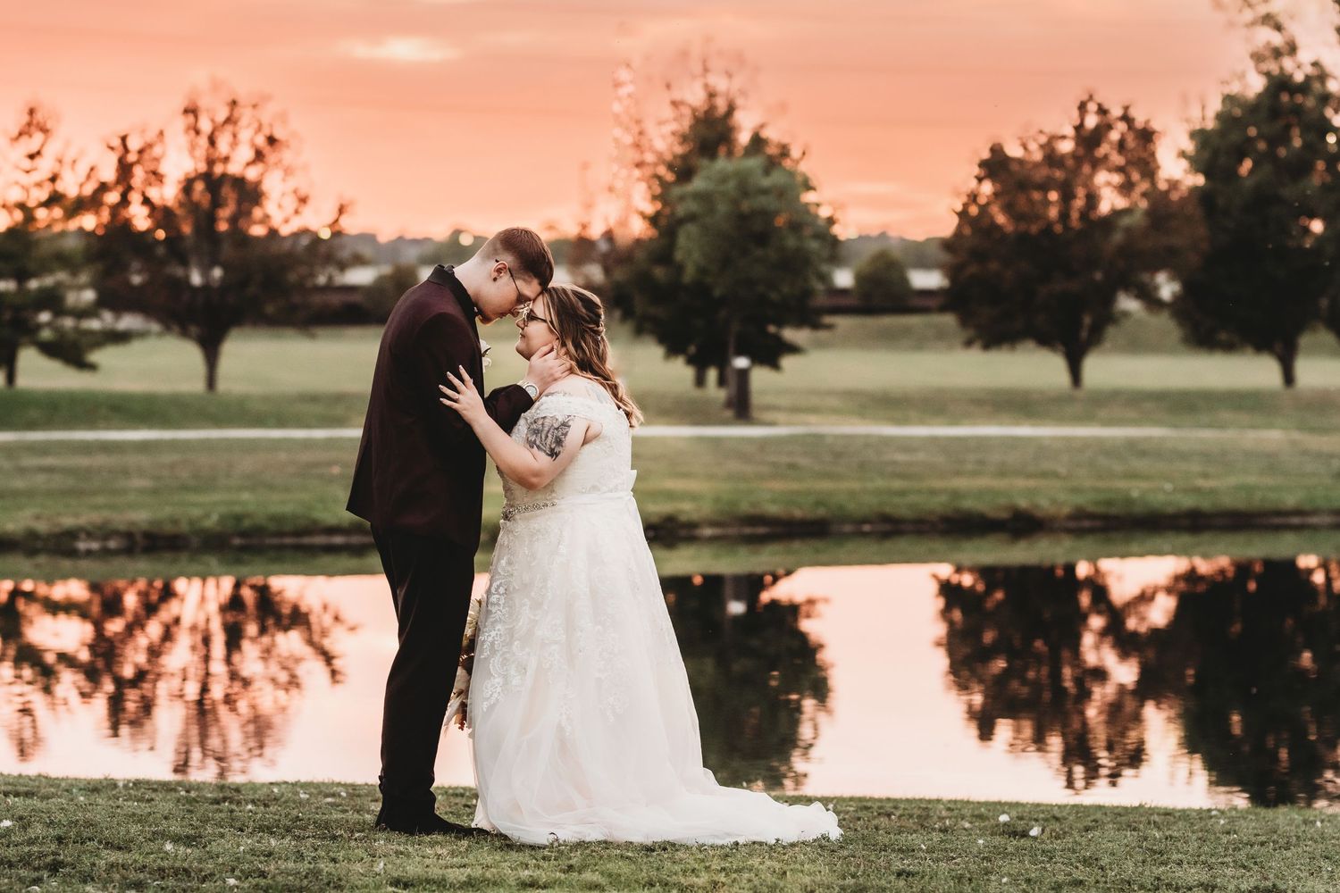 Bride and groom share a romantic kiss at sunset beside a reflective pond at their wedding.