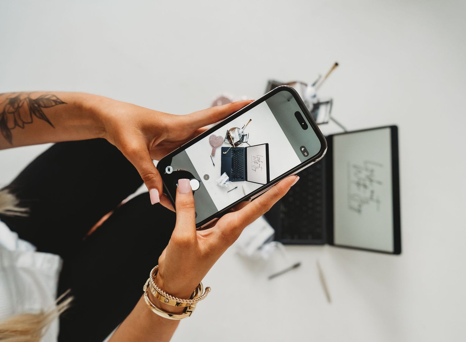 Tattooed woman photographing a flat lay with iPhone above a white desk workspace.