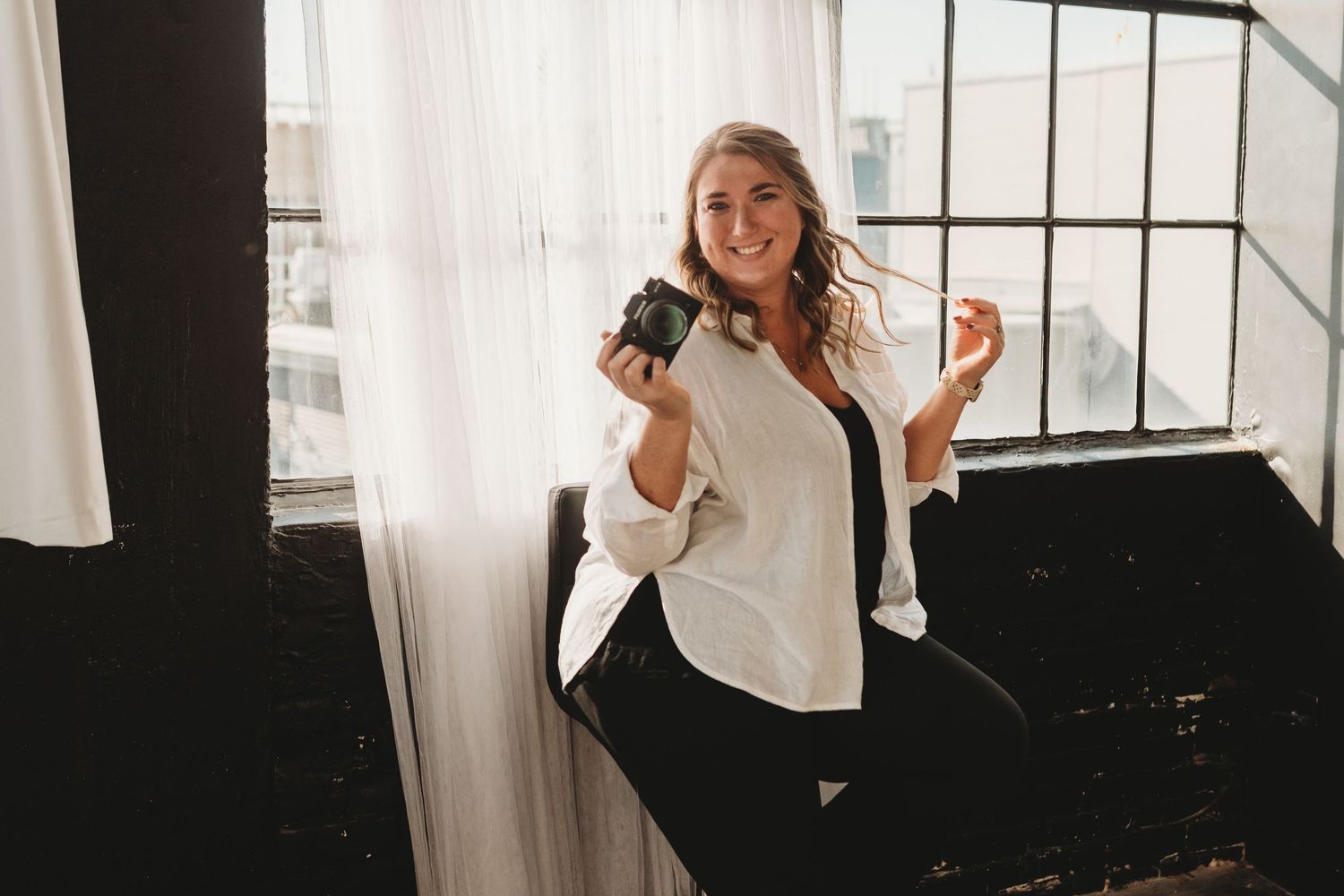 Smiling blonde woman holding a coffee mug seated by large industrial windows in bright studio.