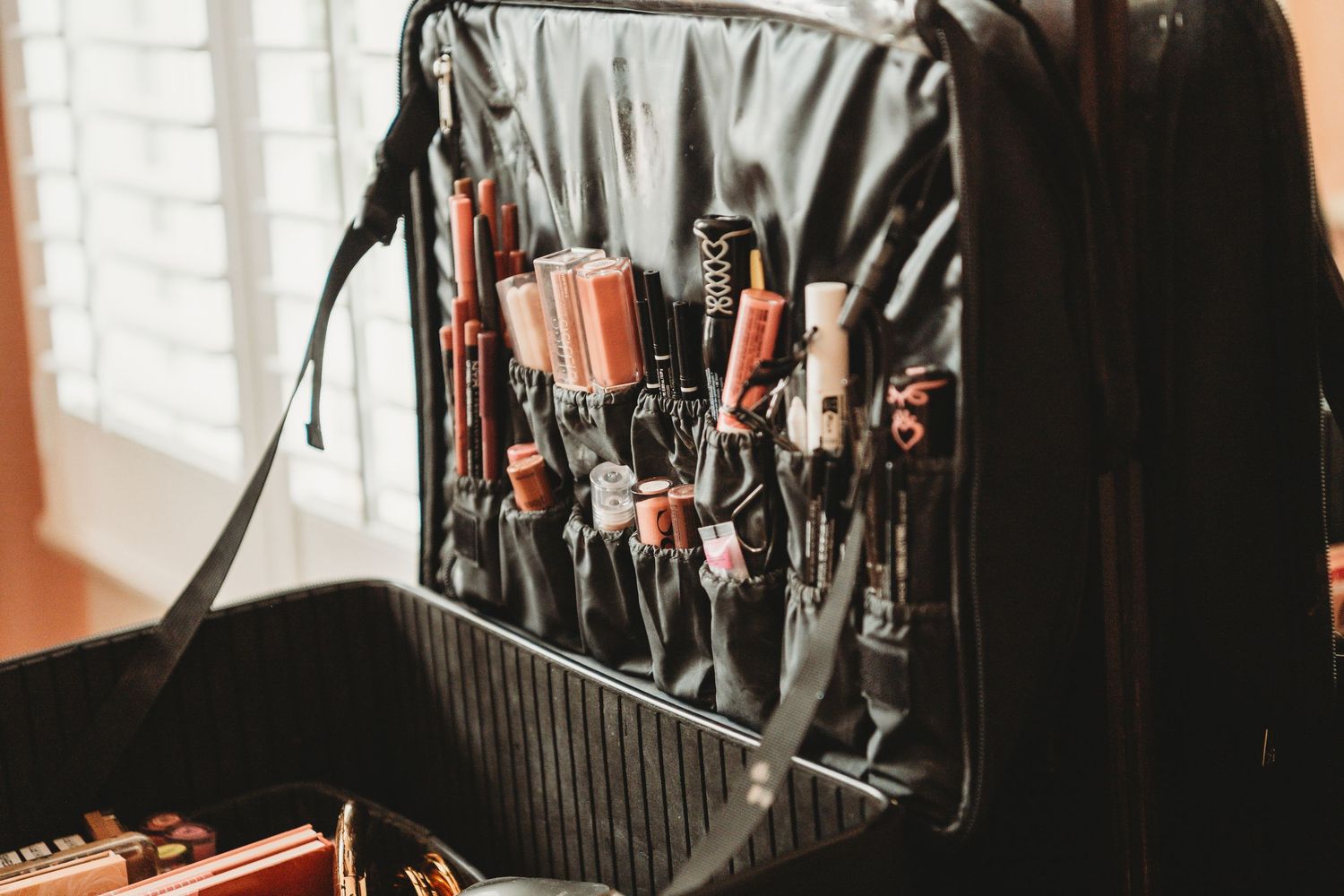 Open makeup artist travel case displaying organized brushes and beauty tools in black fabric pockets near a window.