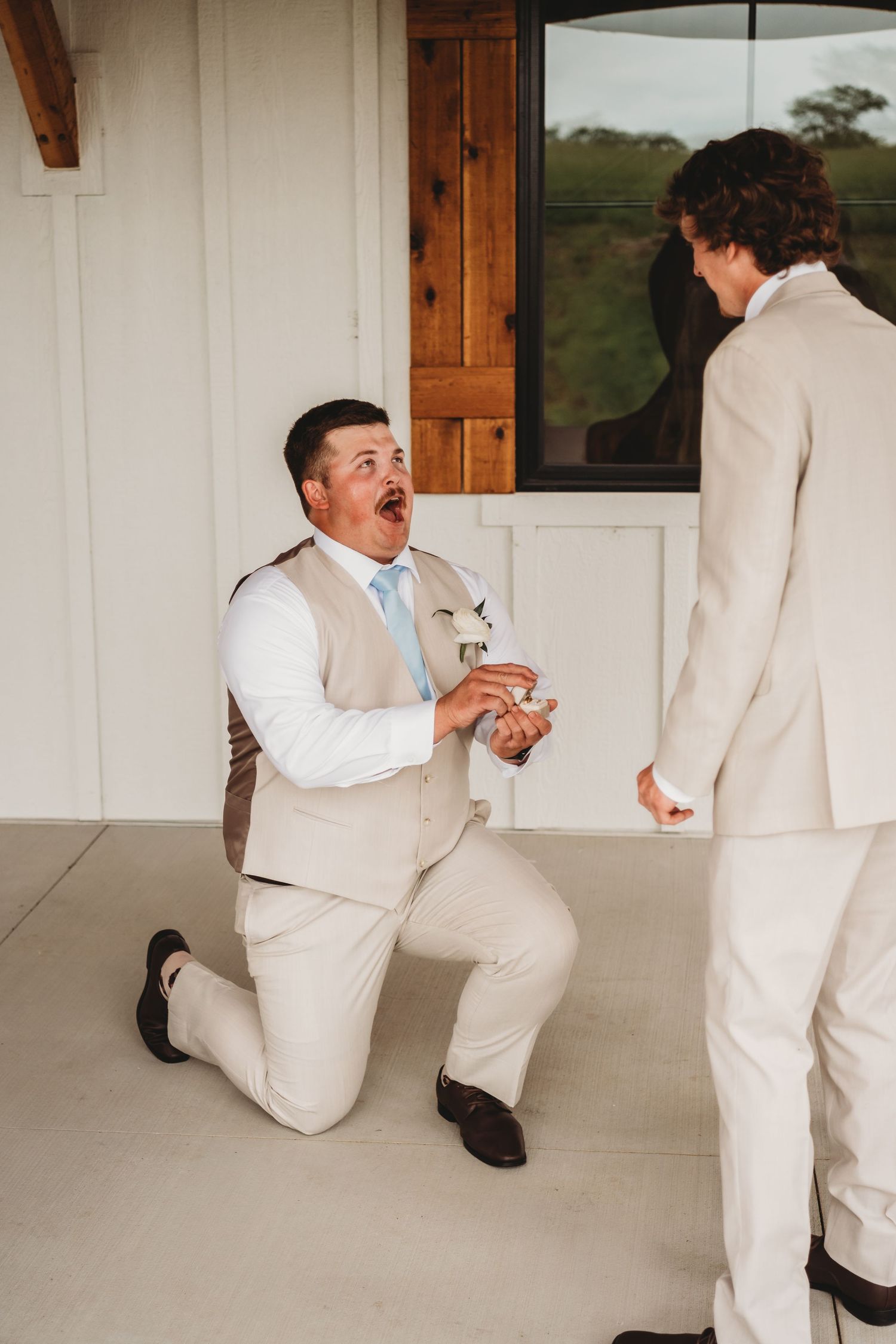 Groomsman in tan suit kneeling dramatically while joking with groom before wedding ceremony.
