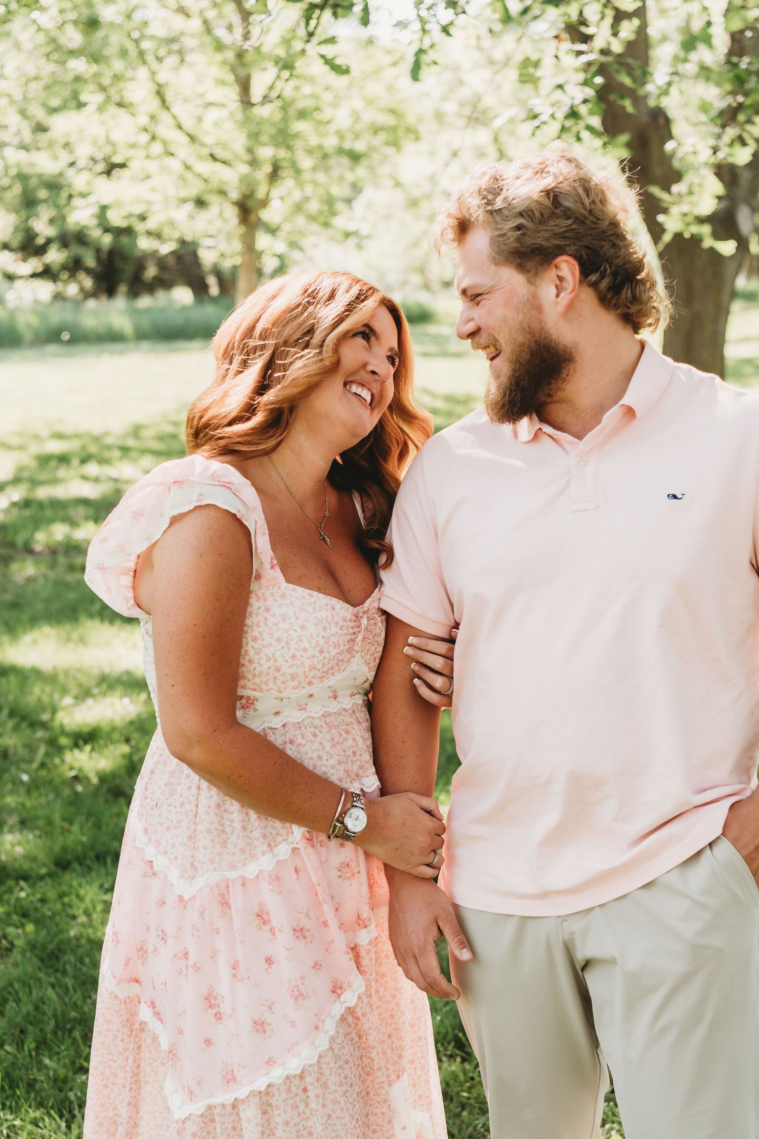 Smiling couple in a lush green park, woman in pink floral dress and man in pink polo shirt.
