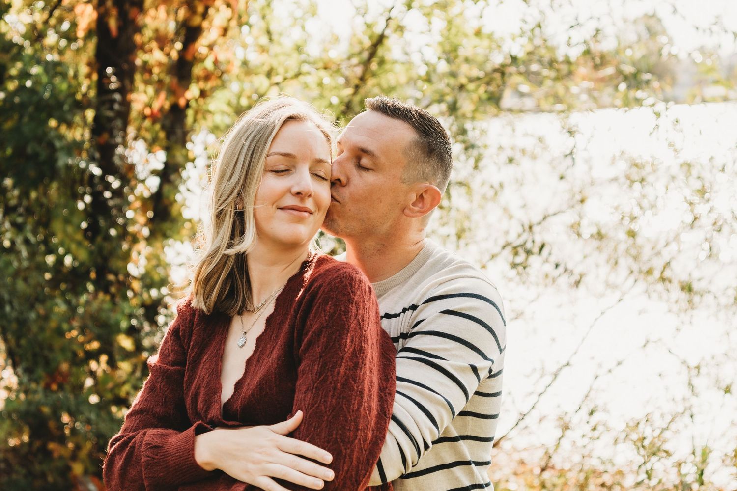 Couple sharing a tender moment outdoors in autumn, man kissing woman's temple near a sunlit tree.