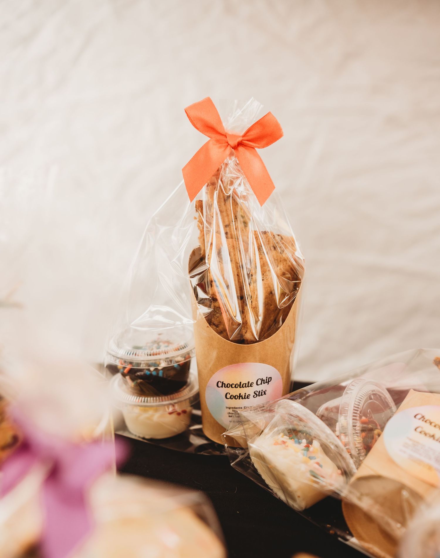Chocolate chip cookie stix gift set in clear bag with orange bow, displayed with assorted baked goods on a table.