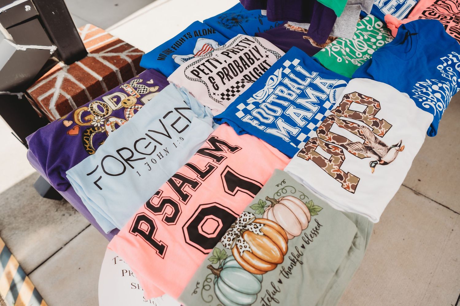 Colorful graphic t-shirts displayed on a table featuring religious, football, and fall-themed designs at an outdoor market.