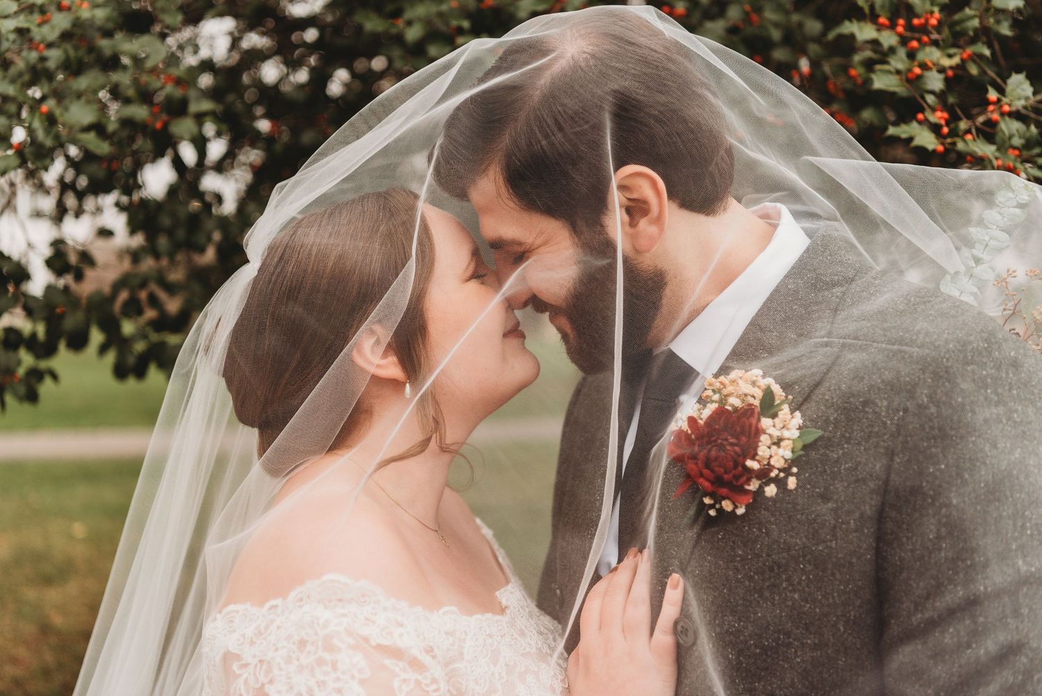 Bride and groom sharing intimate moment under flowing veil with berry trees in background.