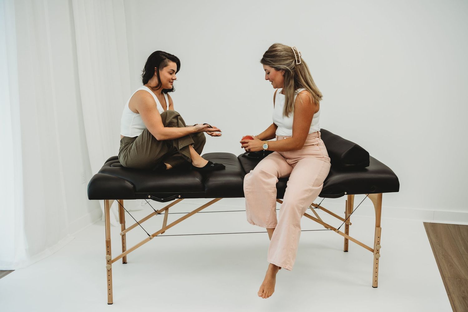 Two women sit on a black massage table in a bright white studio, engaged in a consultation session.