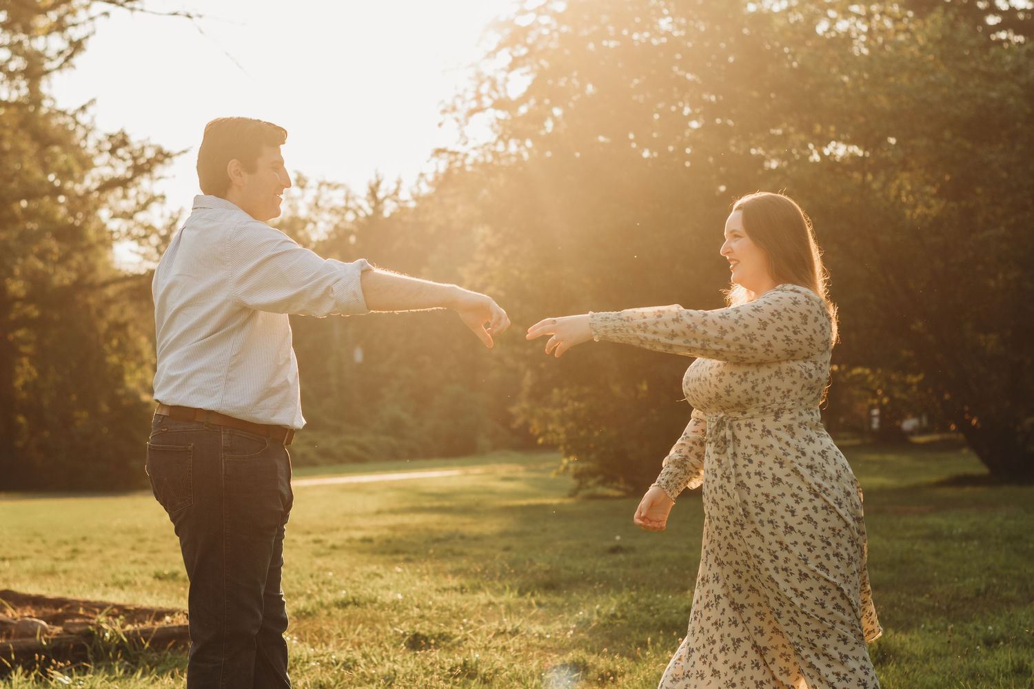 A couple reaches toward each other playfully in a glowing golden sunset park setting.