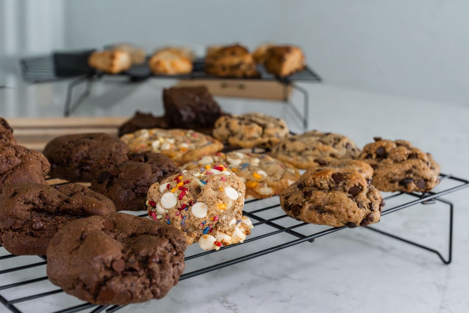 Assorted freshly baked cookies on wire cooling racks, including chocolate, M&M, and chocolate chip varieties.