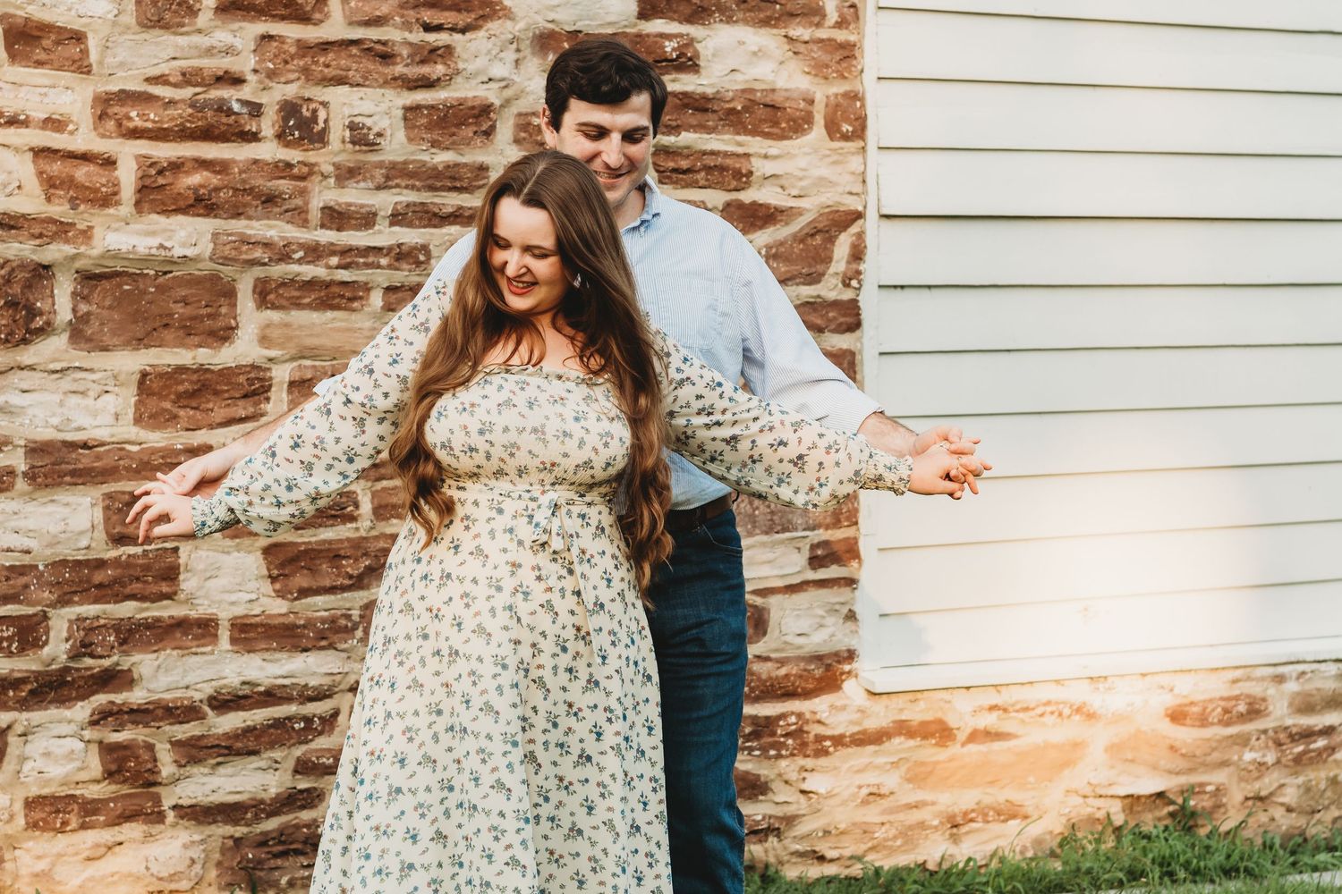 Couple dancing and laughing together outdoors against a rustic stone wall during engagement session.
