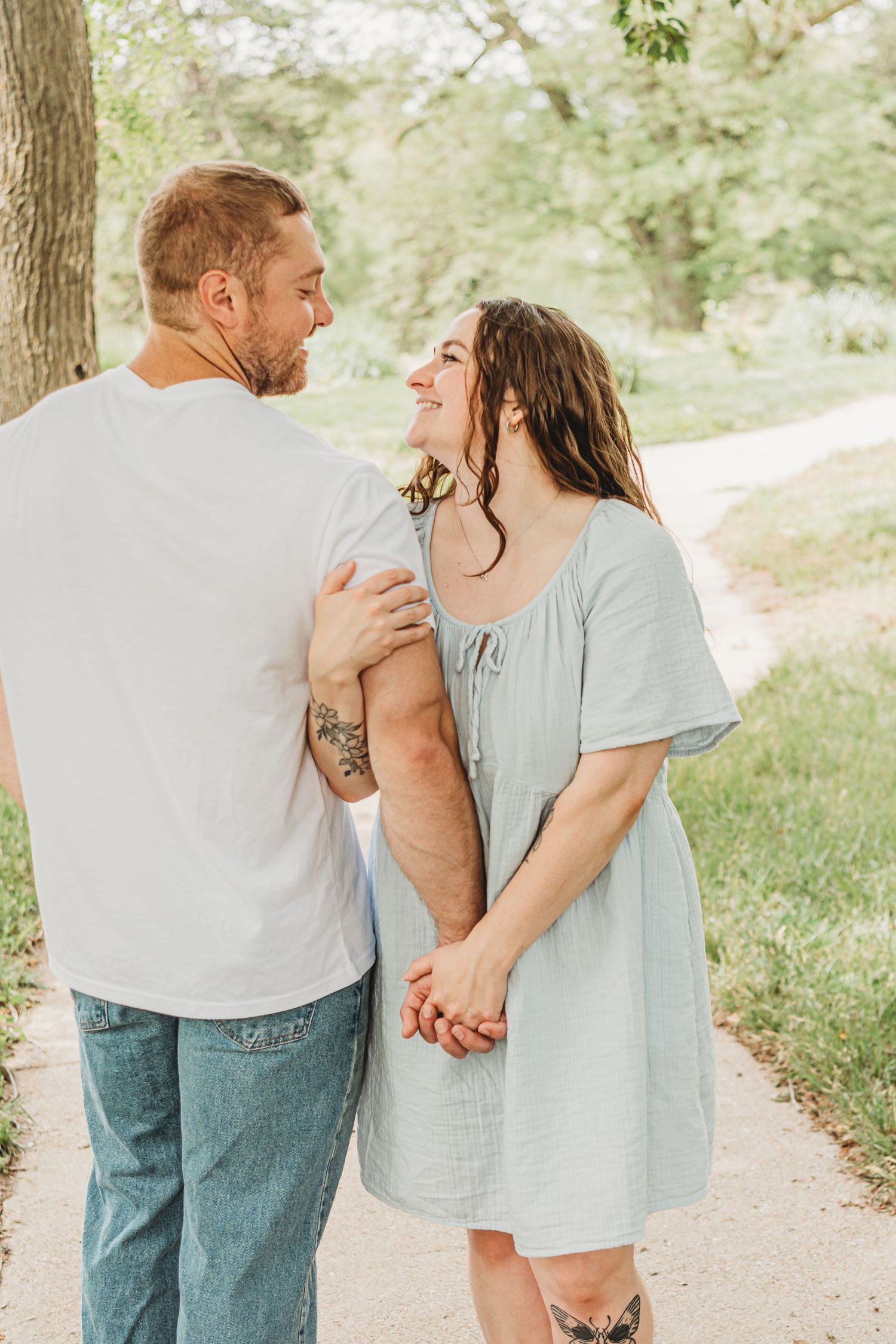 A couple holds hands and smiles at each other while walking along a sunny tree-lined path.