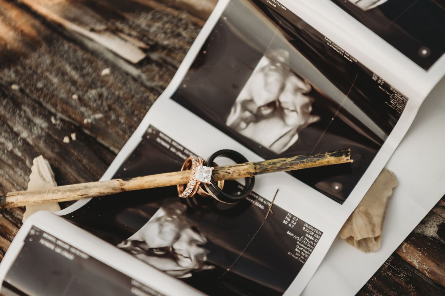 Wedding rings and a rustic twig rest on black and white photographs on a wooden surface.