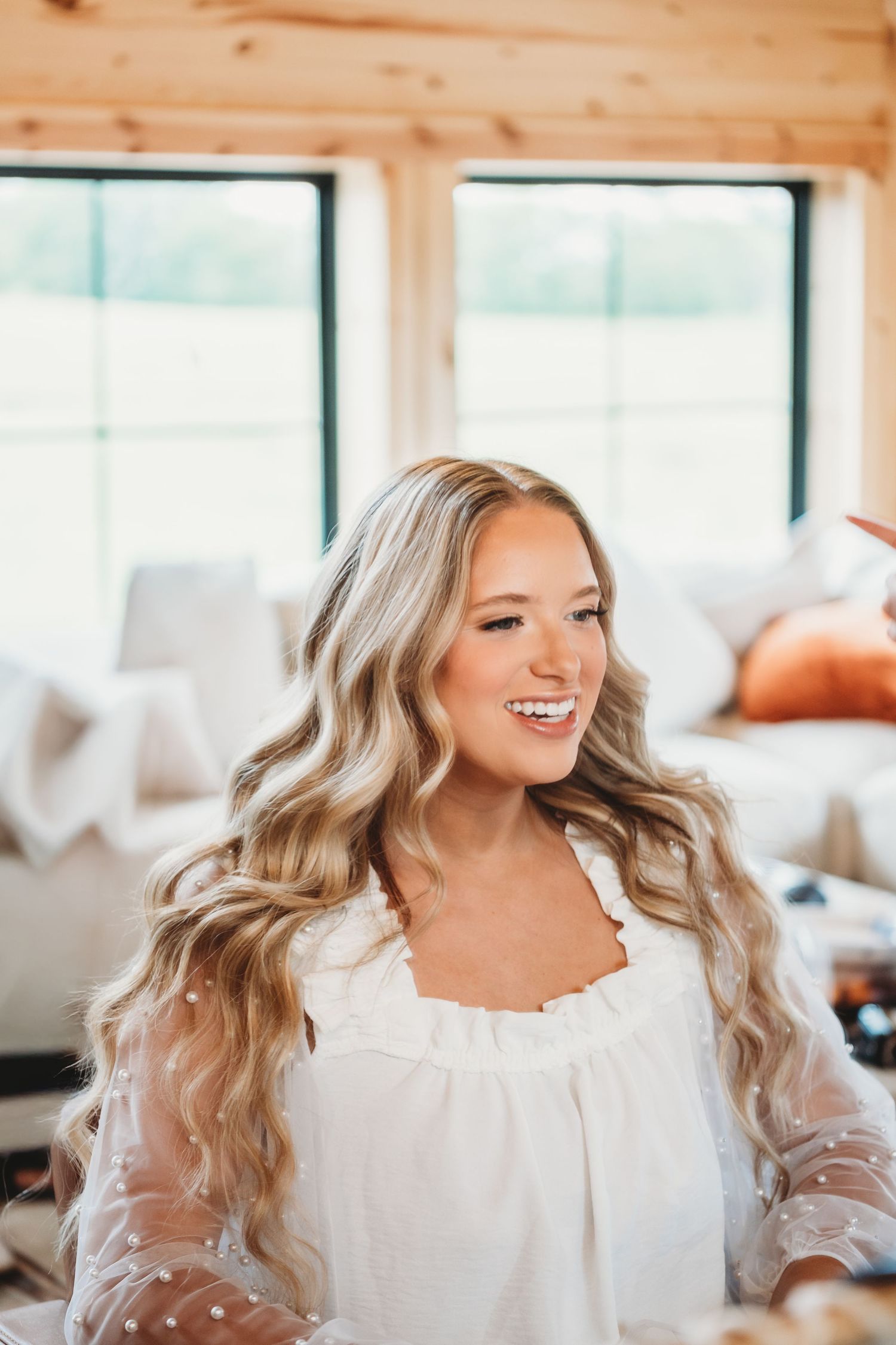 Smiling blonde bride in white dress with wavy hair getting ready in a bright barn venue.