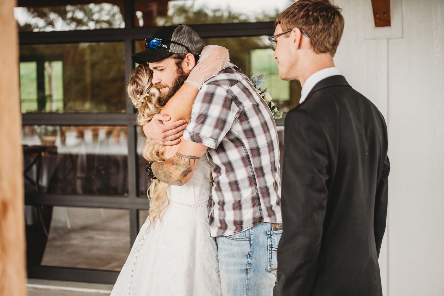 Bride in white dress sharing an emotional hug with a guest in a plaid shirt before ceremony.
