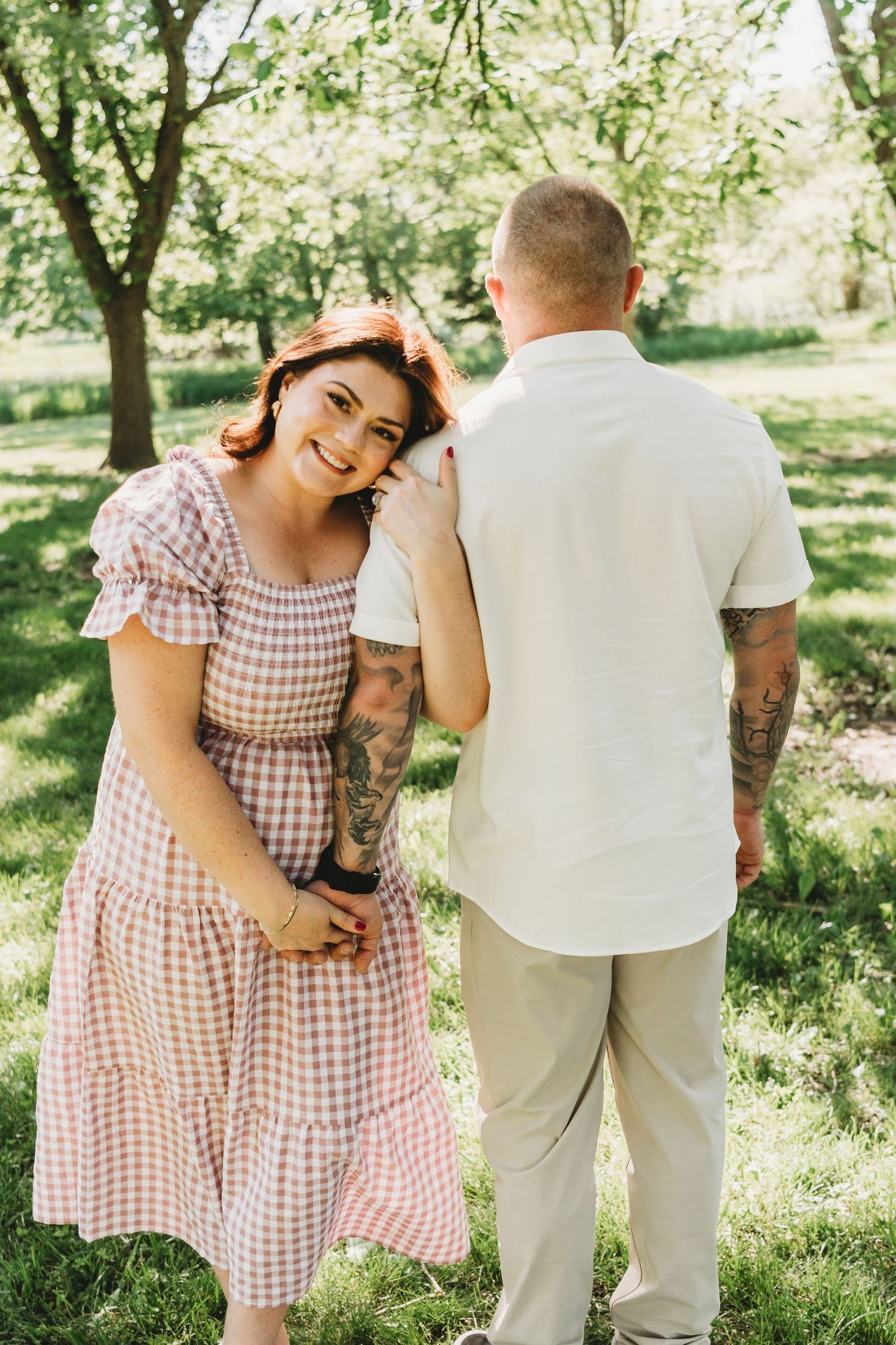 A smiling tattooed woman in a pink gingham dress holds hands with her partner outdoors.