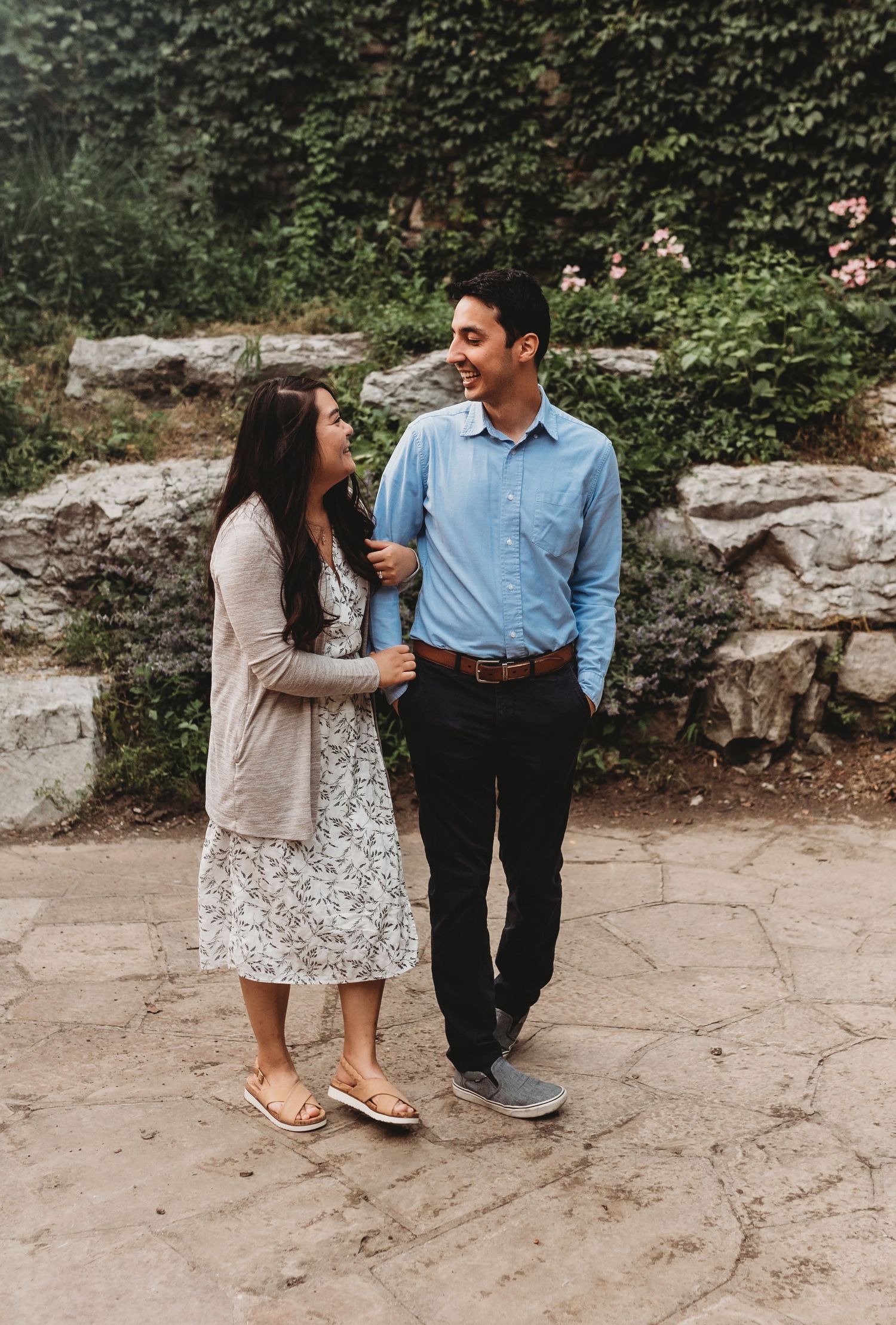 A couple laughs together while walking through a stone garden surrounded by lush greenery.