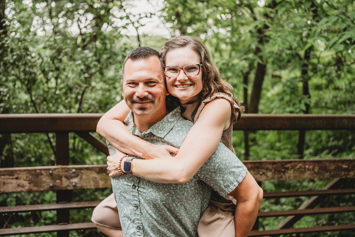 Happy couple embracing on wooden bridge surrounded by lush green trees.