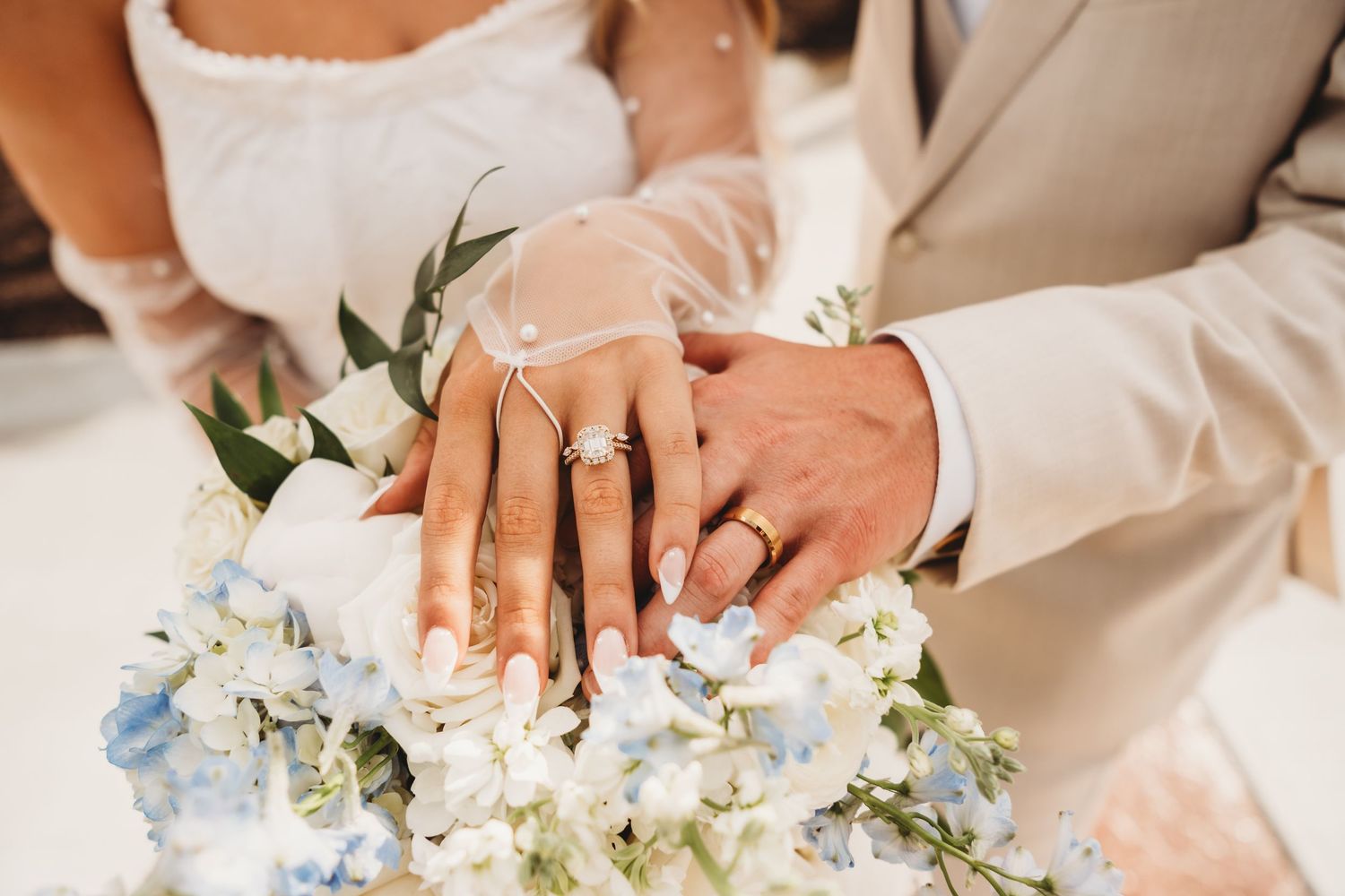 Close-up of newlyweds hands displaying wedding rings resting on a blue and white floral bouquet.
