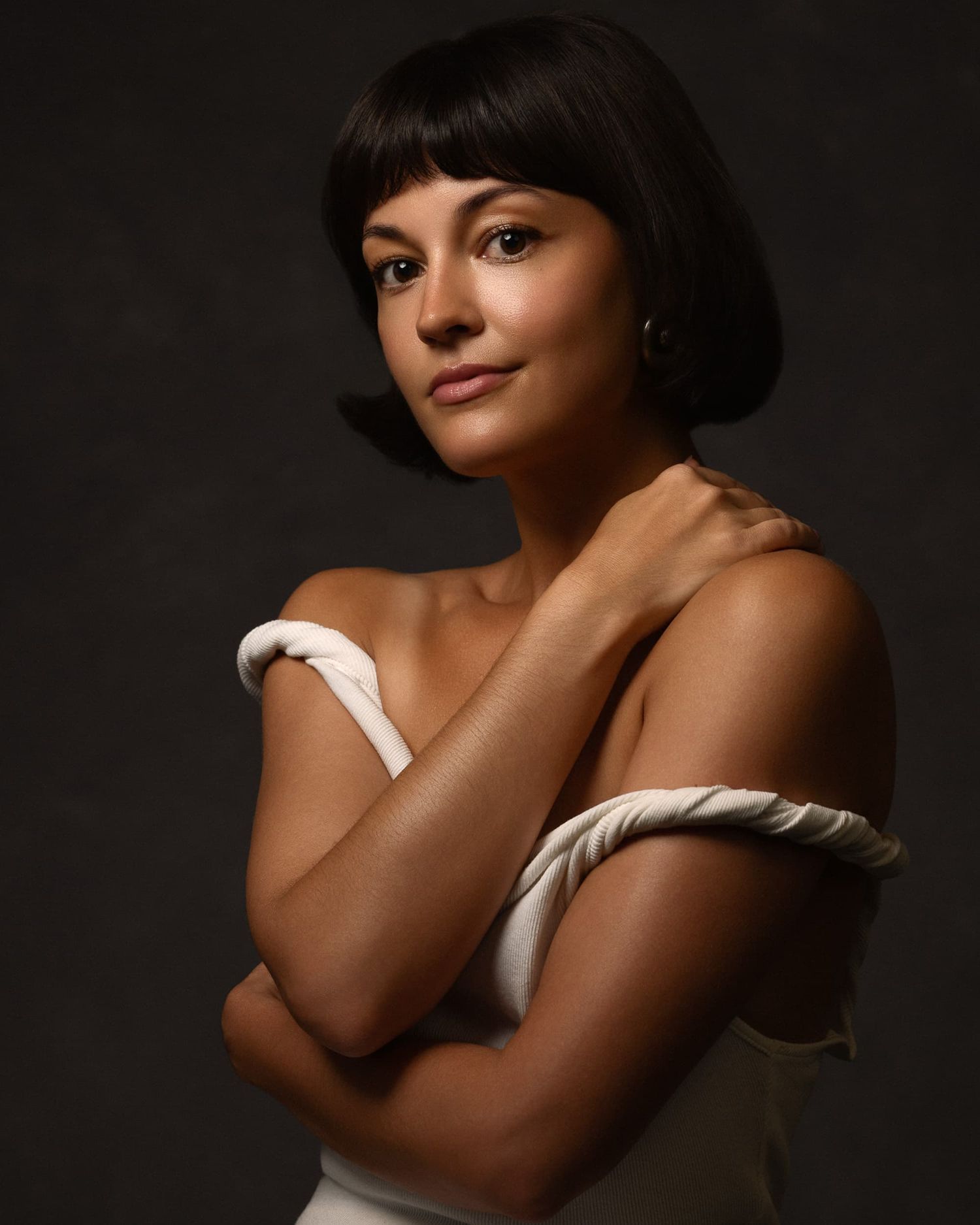 Woman with bob haircut in elegant off-shoulder white dress, arms crossed, posing against dark background.