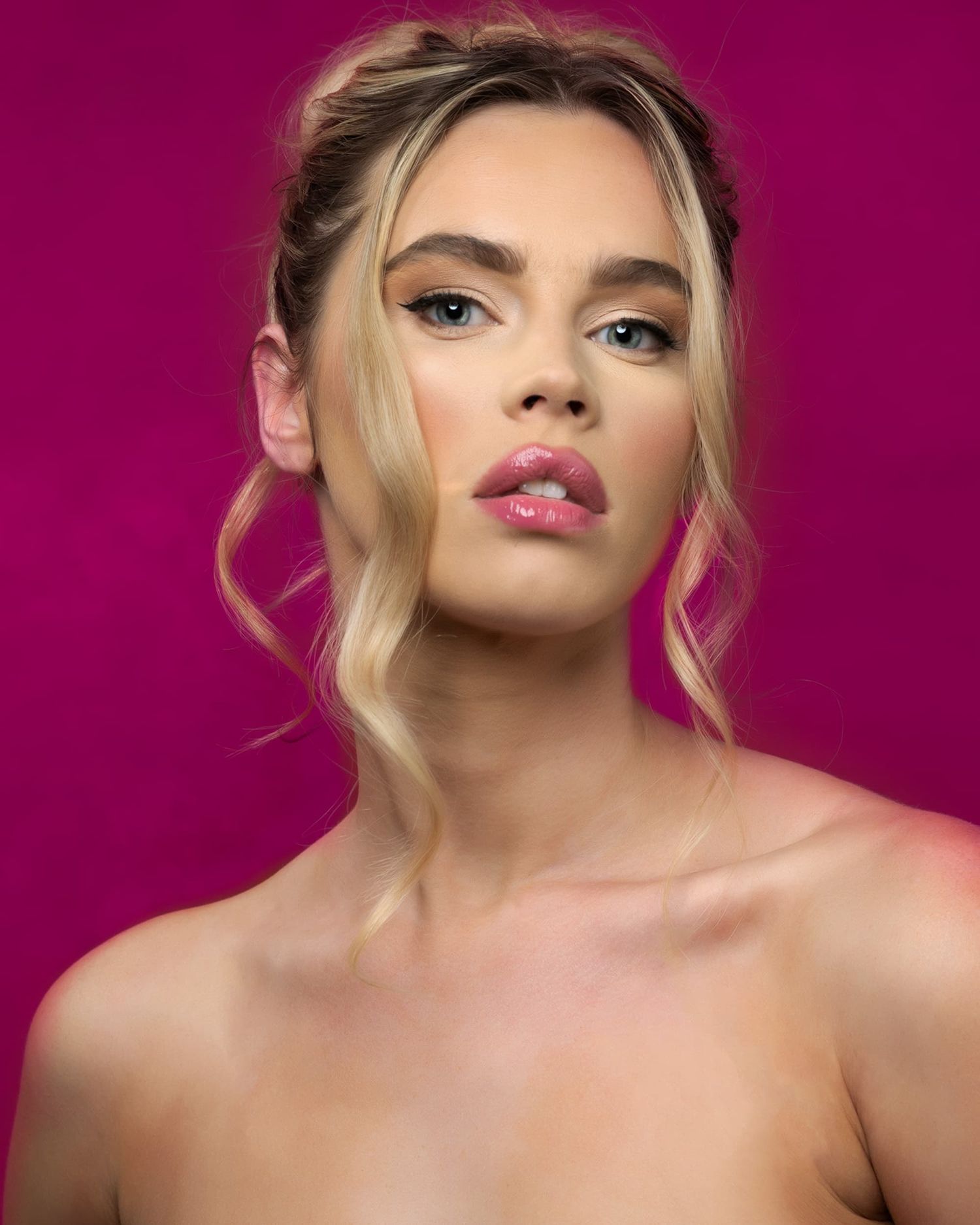 Blonde woman with pink glossy lips and elegant updo poses against a vibrant magenta background with flawless makeup.