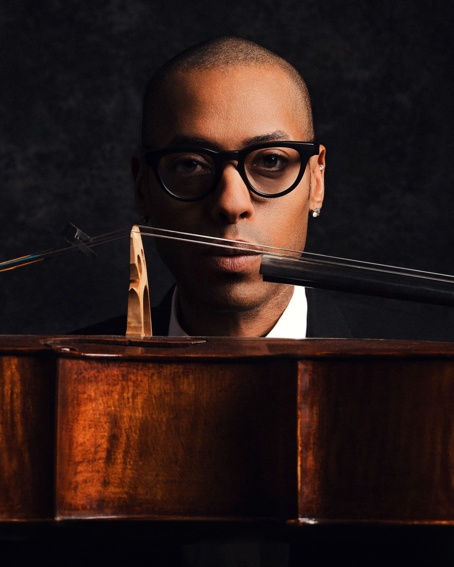 Close-up portrait of a young man with glasses holding a cello bow, peering over the instrument.