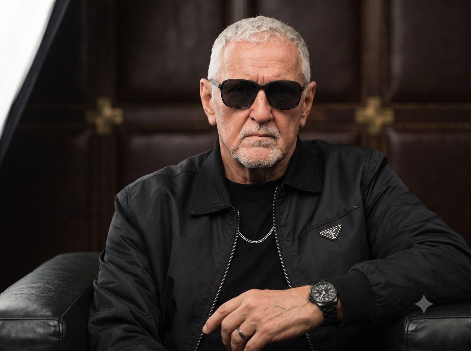 Older man with gray hair wearing sunglasses and black jacket seated on leather sofa.