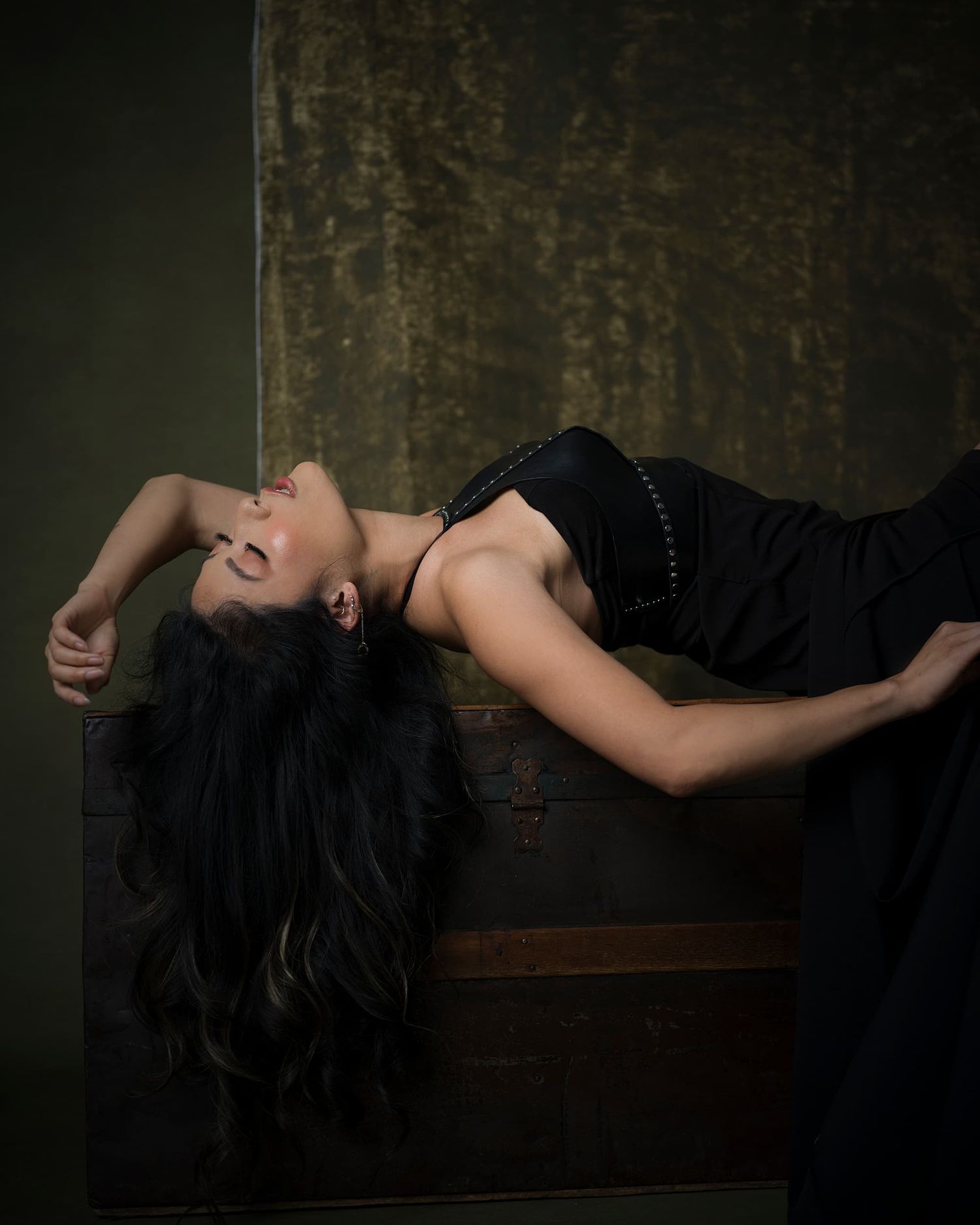 Woman in black gown reclines gracefully on a vintage trunk, head tilted back.