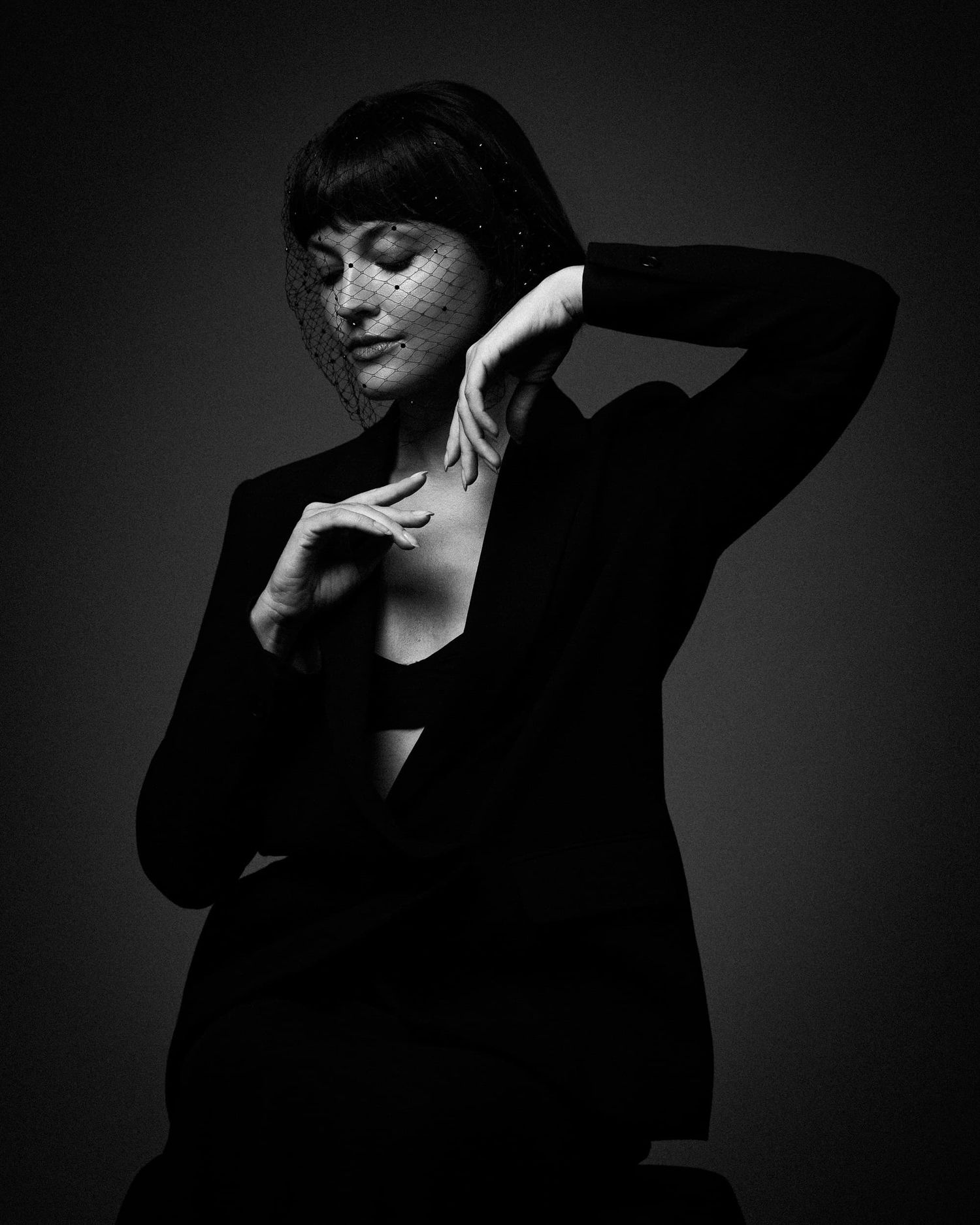 Dramatic black and white photo of a woman in a black blazer wearing a veil fascinator hat.