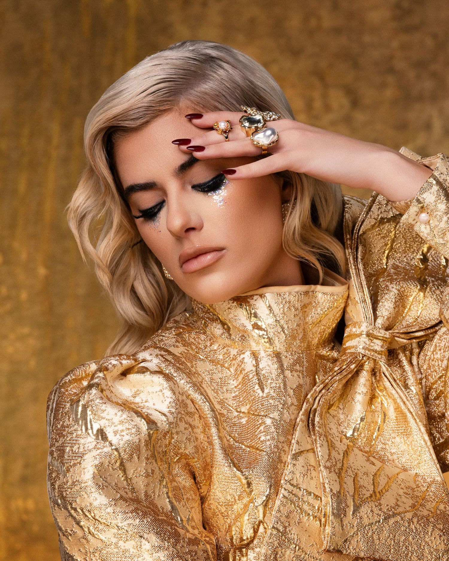 Blonde model in a gold brocade dress showcasing bold rings and dark nail polish.