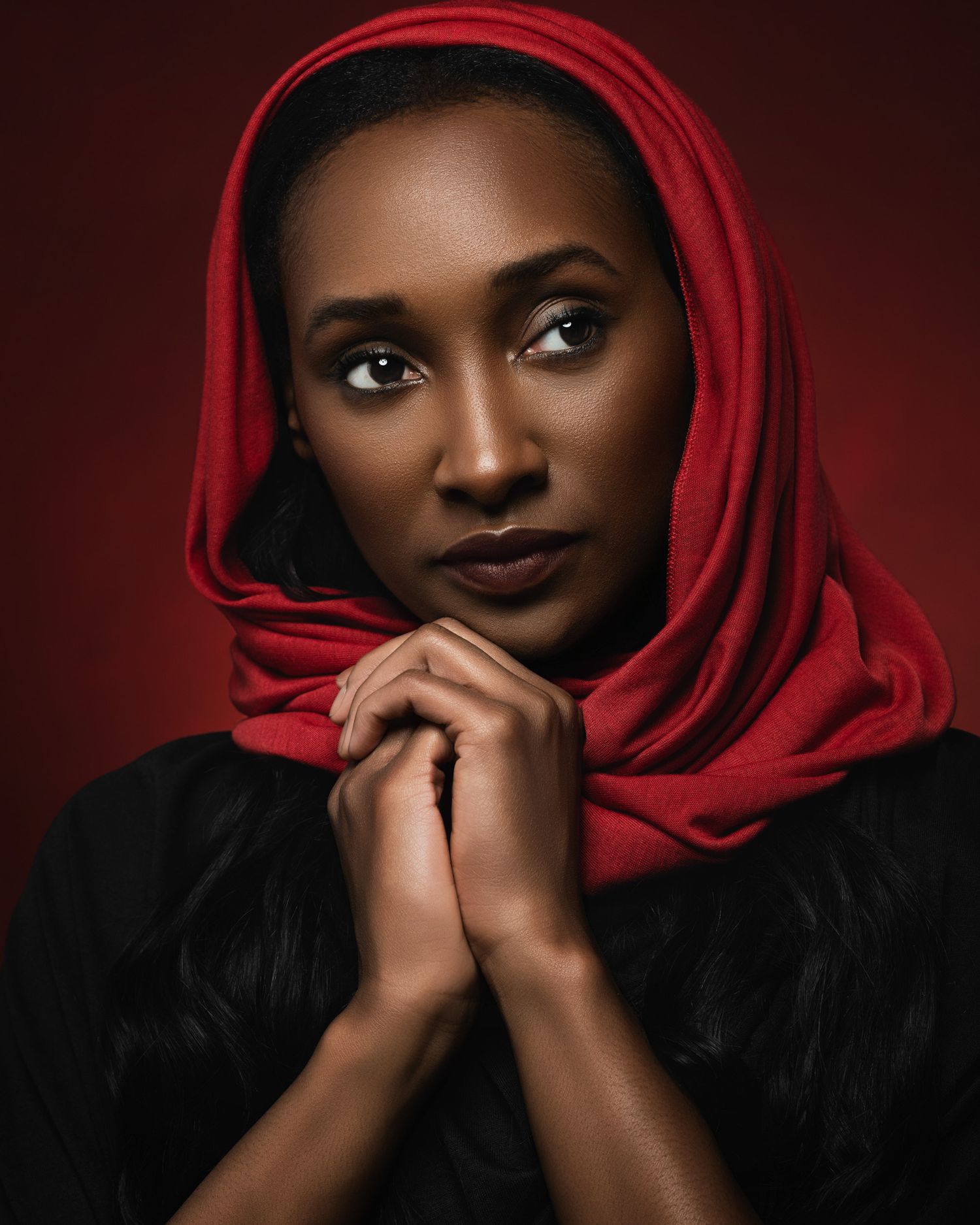 A Black woman wearing a red hijab clasps her hands together against a deep red background.