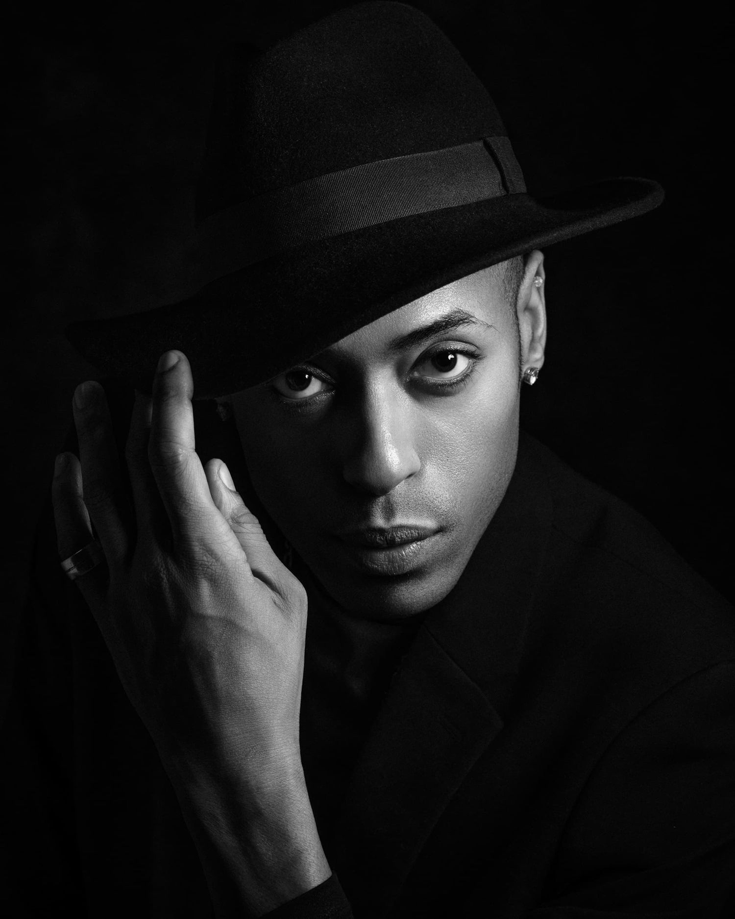 Black and white portrait of a young man wearing a wide-brim fedora hat, posing with hand raised to face.