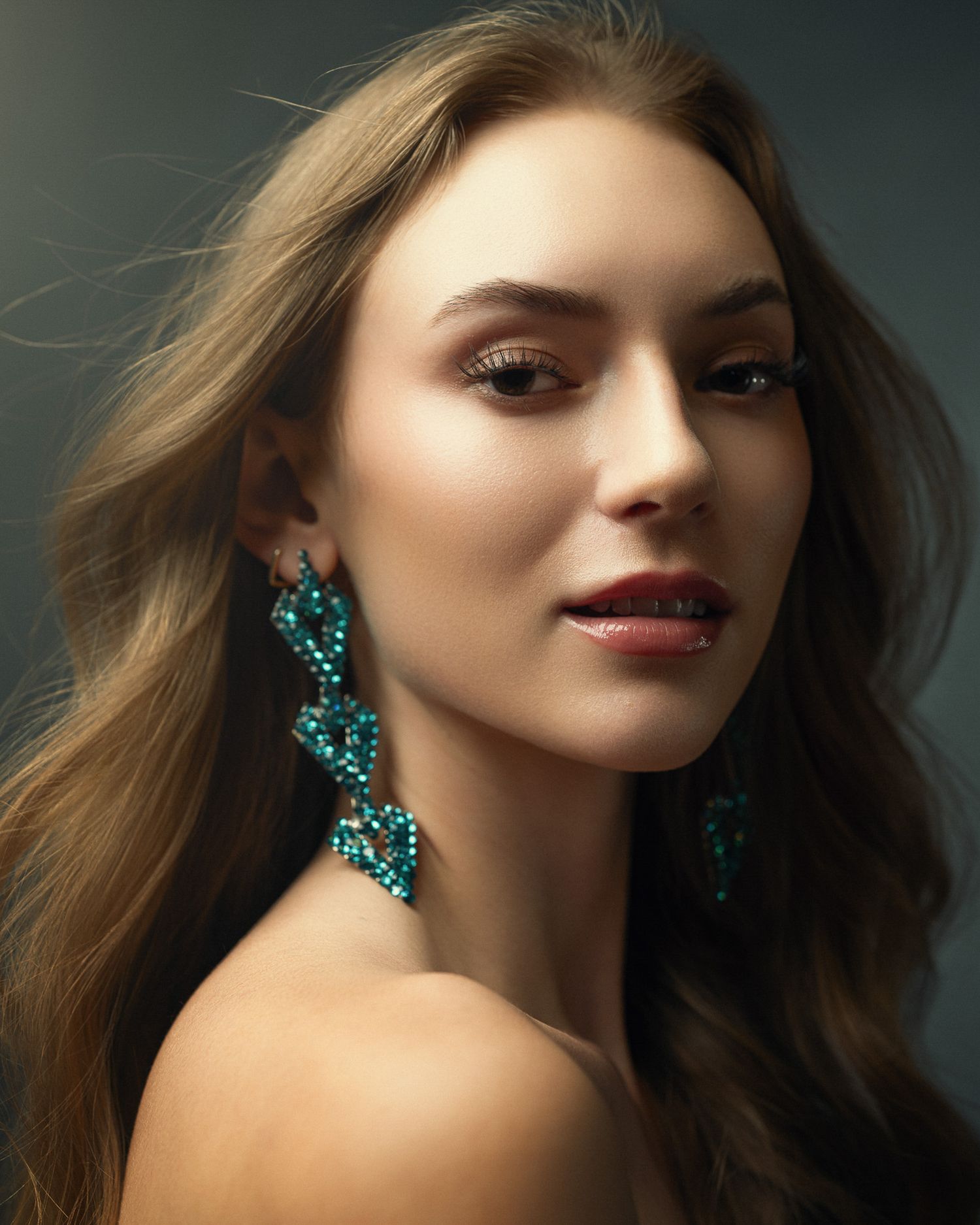 Close-up portrait of woman wearing long turquoise crystal drop earrings against gray background.
