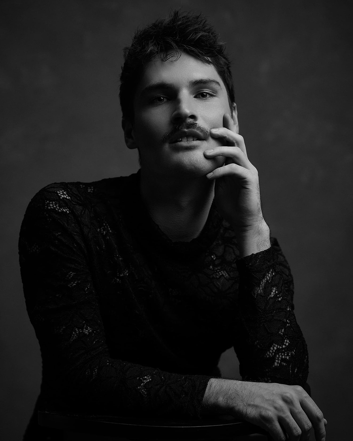 Black and white portrait of a young man with a mustache wearing a black lace top in a studio.