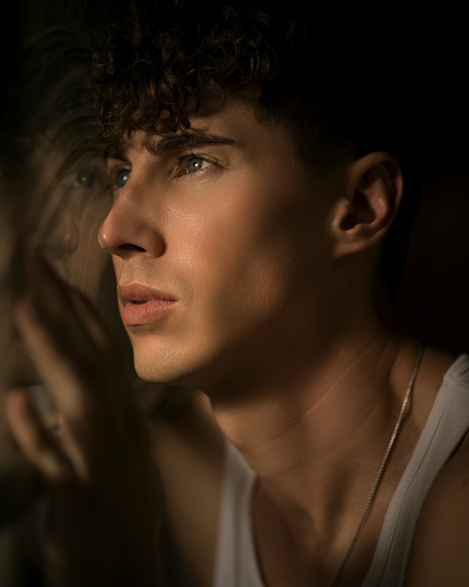 Young man with curly hair and gold necklace gazes pensively through glass in dramatic moody lighting.