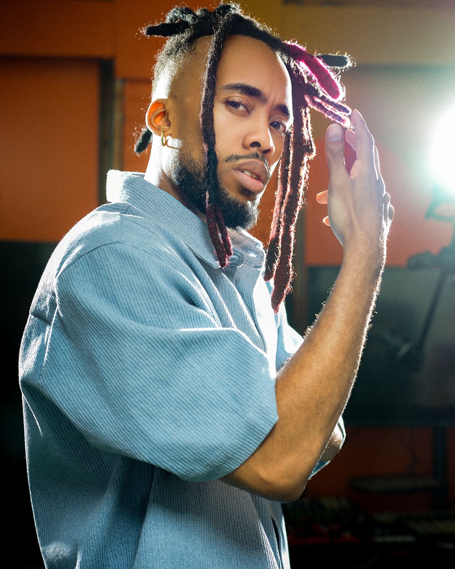 Young man with colorful dreadlocks wearing a light blue corduroy shirt posing in a warmly lit room.