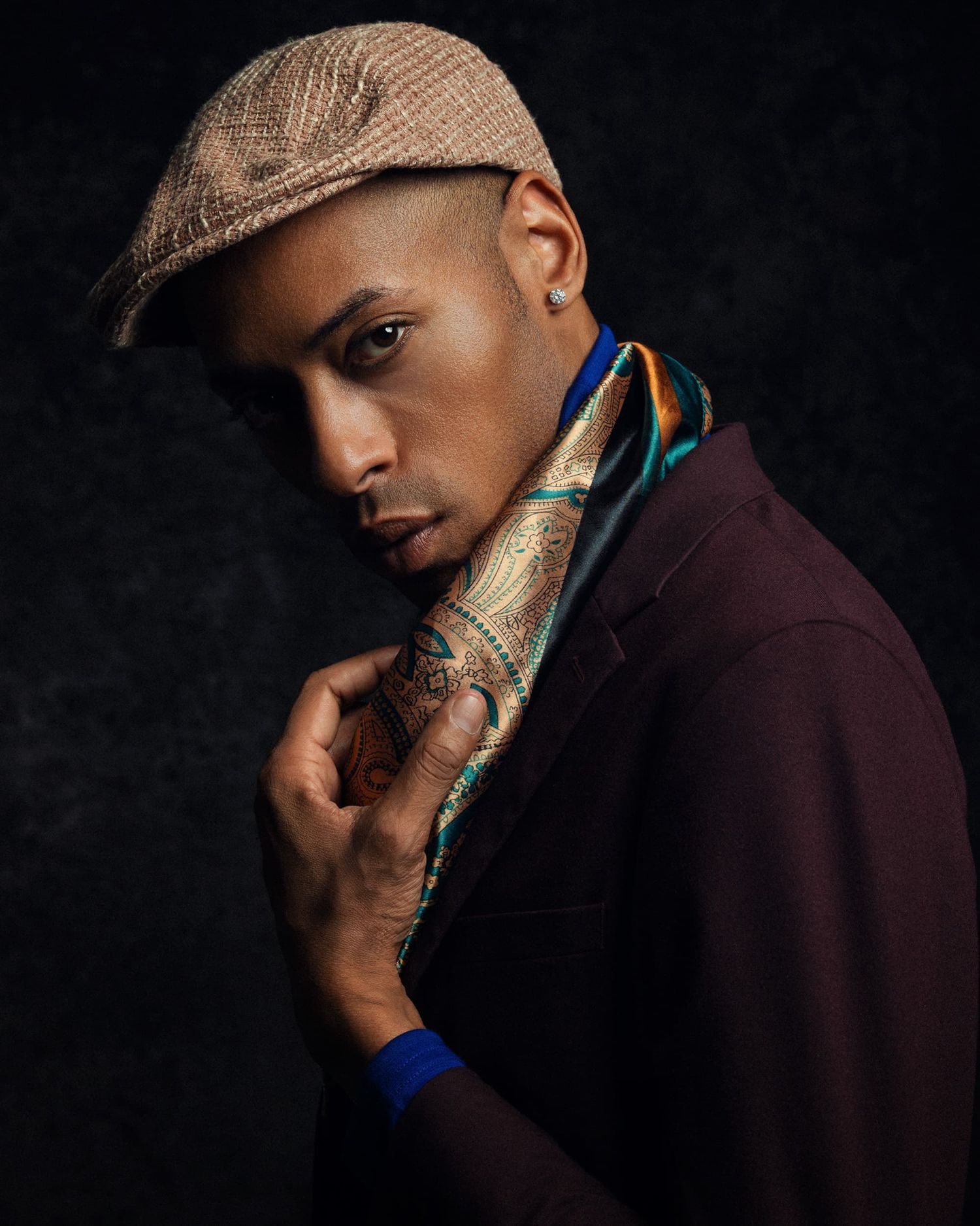 Stylish young man wearing a flat cap and colorful scarf with a burgundy blazer in a dramatic portrait.